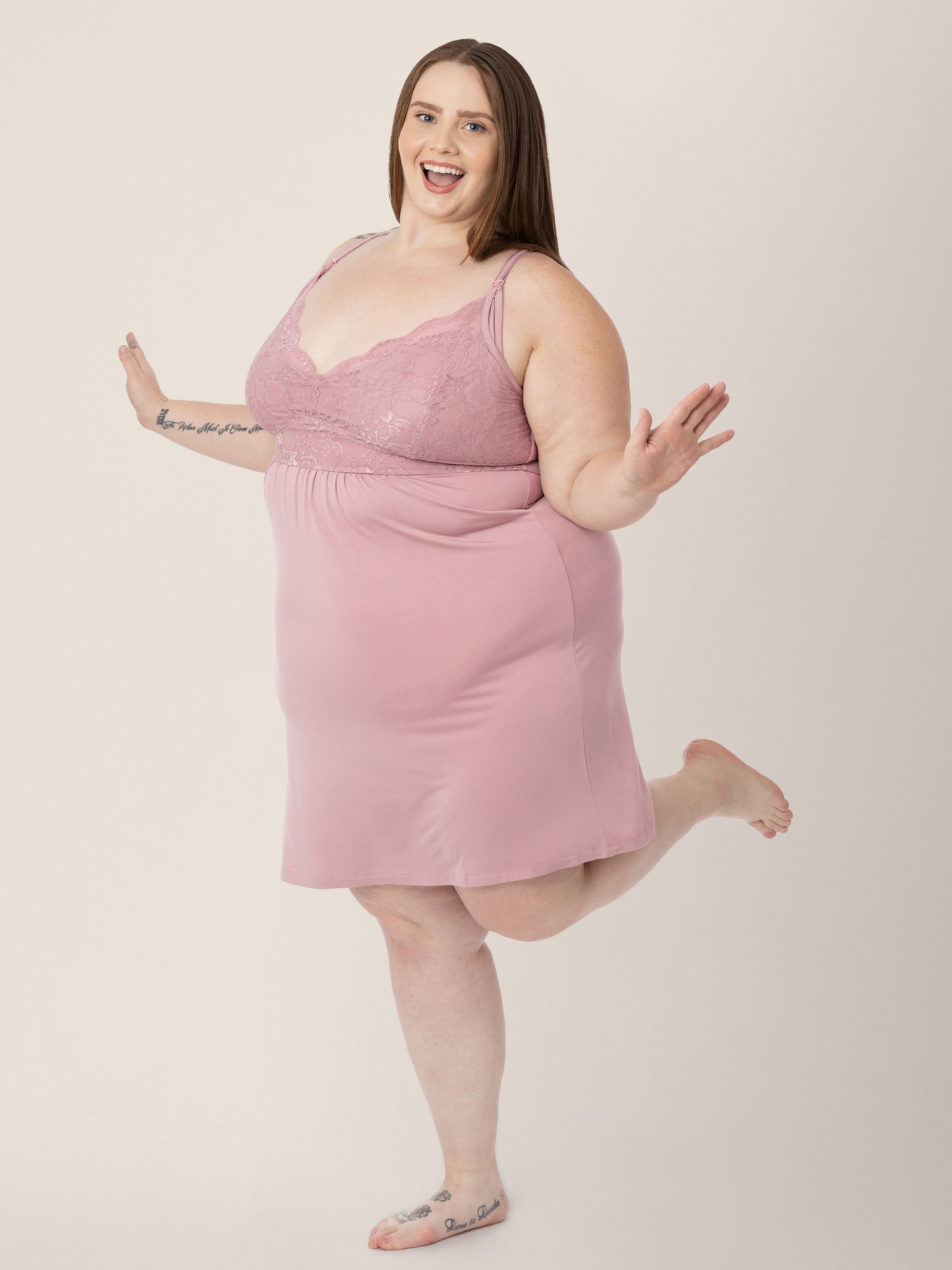 Model wearing the Lucille Maternity & Nursing Nightgown in Vintage Pink standing on one leg and smiling. @model_info:Hayley is 5'7" and wearing a 1X.