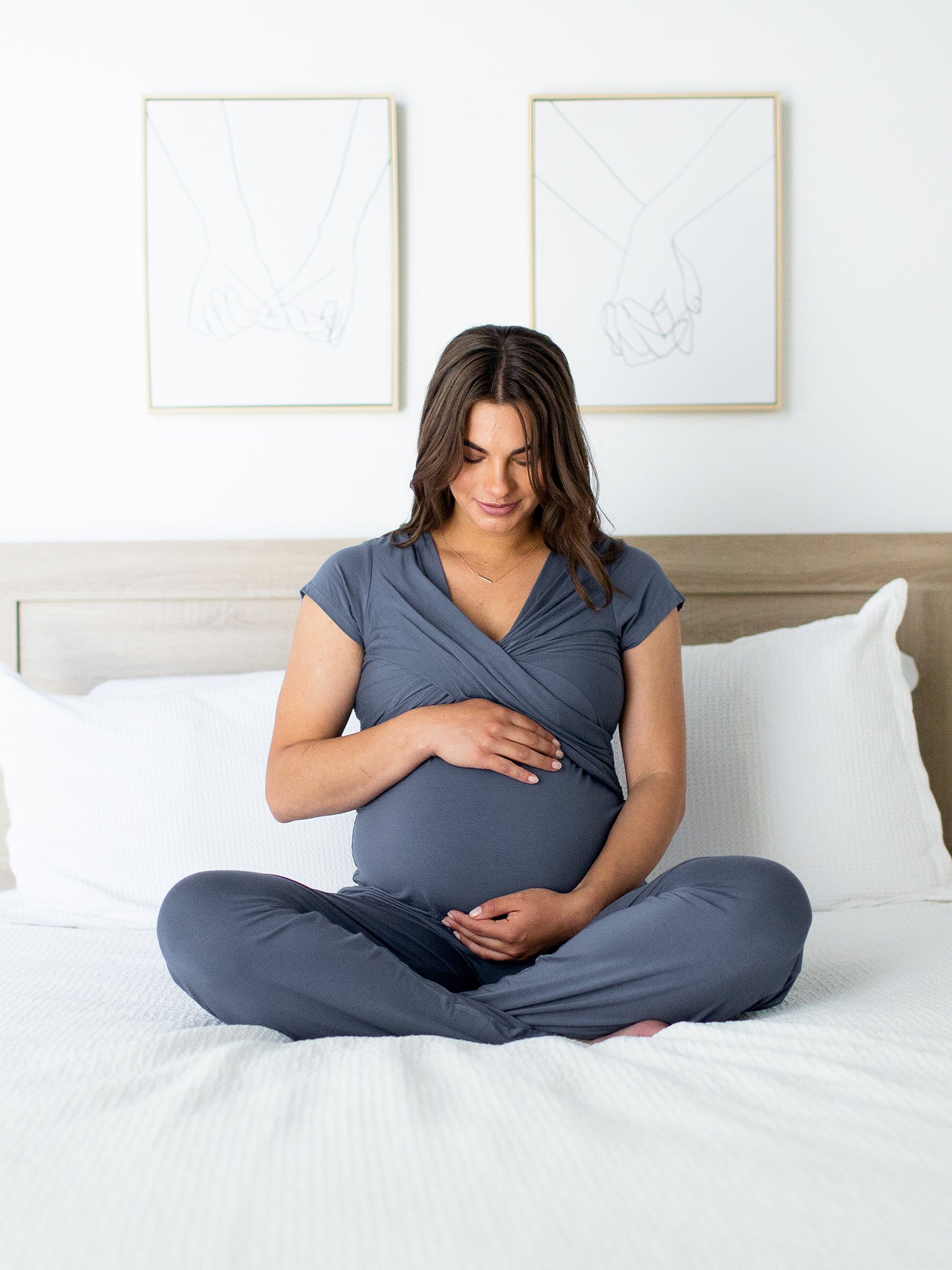 Model sitting on a bed wearing the Davy Maternity & Nursing Pajama Set in Slate Blue holding her pregnant belly sitting on a white bed. @model_info:Rayne is wearing a Medium.