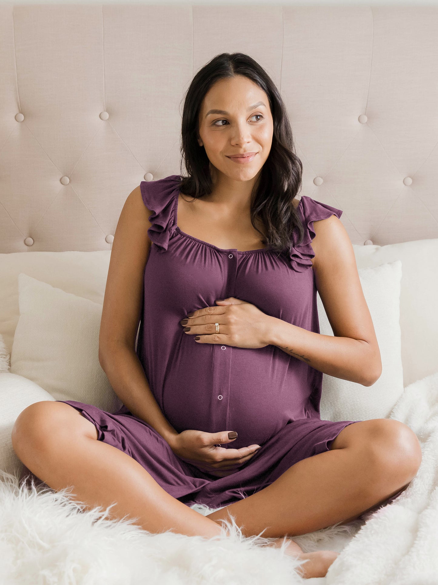 Model sitting on a bed while wearing the Ruffle Strap Labor & Delivery Gown in Burgundy Plum with her hands on her pregnant belly. @model_info:Julana is 5'9" and wearing an XS/S.