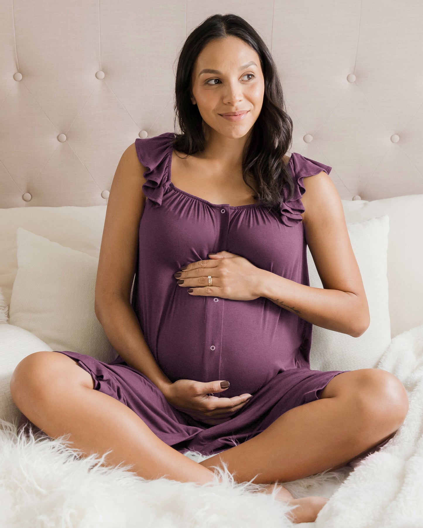 Model sitting on a bed while wearing the Ruffle Strap Labor & Delivery Gown in Burgundy Plum with her hands on her pregnant belly. @model_info:Julana is 5'9" and wearing an XS/S.