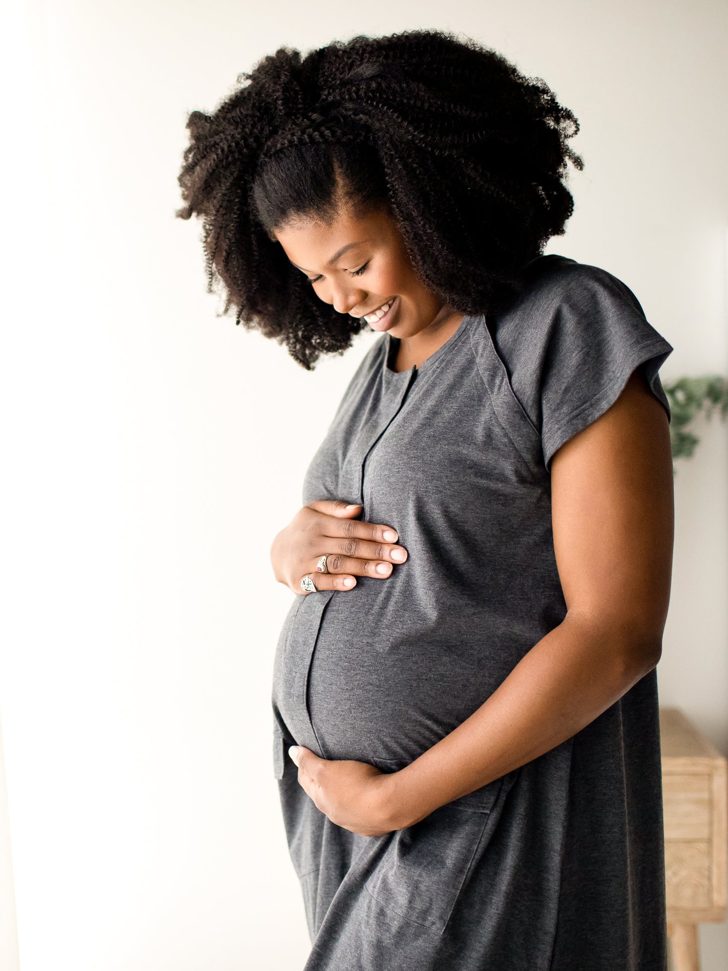 Pregnant model wearing the Universal Labor & Delivery Gown in Grey Heather holding her pregnant belly in a white room. @model_info:Joy is wearing a S/M/L.