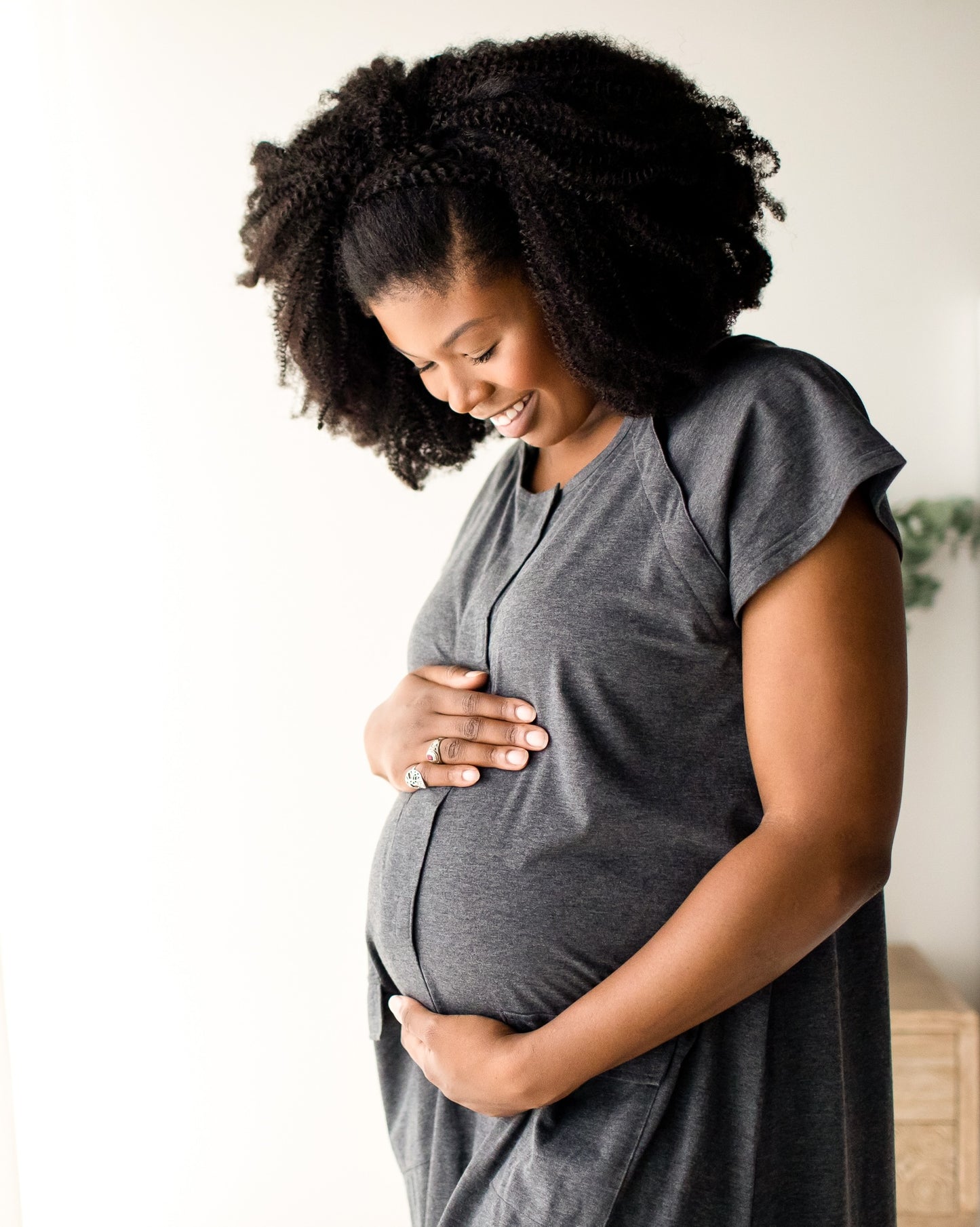 Pregnant model wearing the Universal Labor & Delivery Gown in Grey Heather holding her pregnant belly in a white room. @model_info:Joy is wearing a S/M/L.