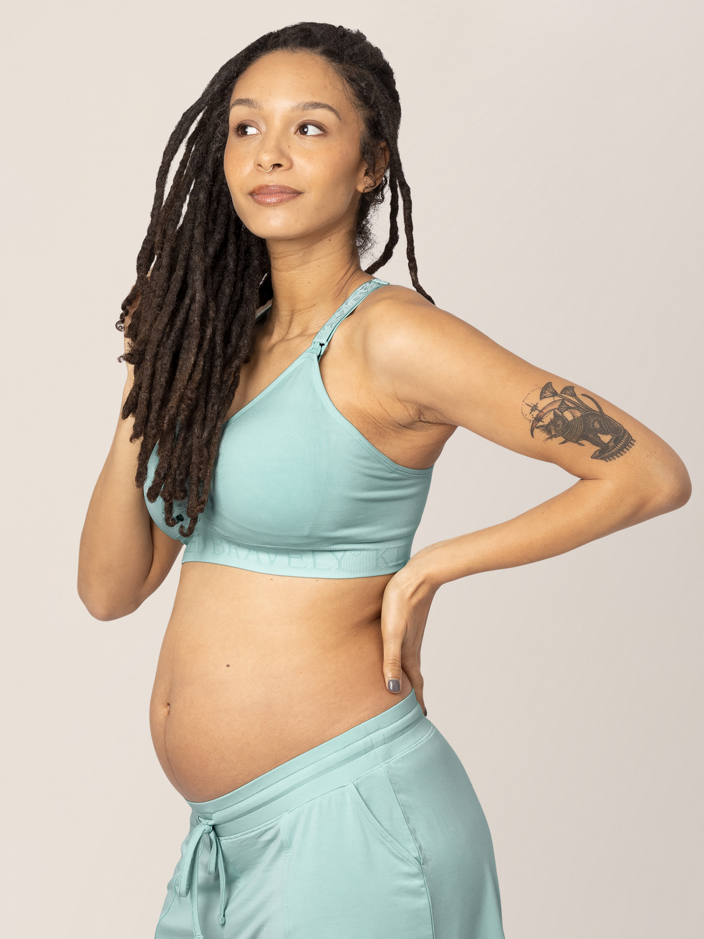 Pregnant model with her hand on her hip and in her hair wearing the Sublime® Hands-Free Pumping & Nursing Sports Bra in Dusty Blue Green.