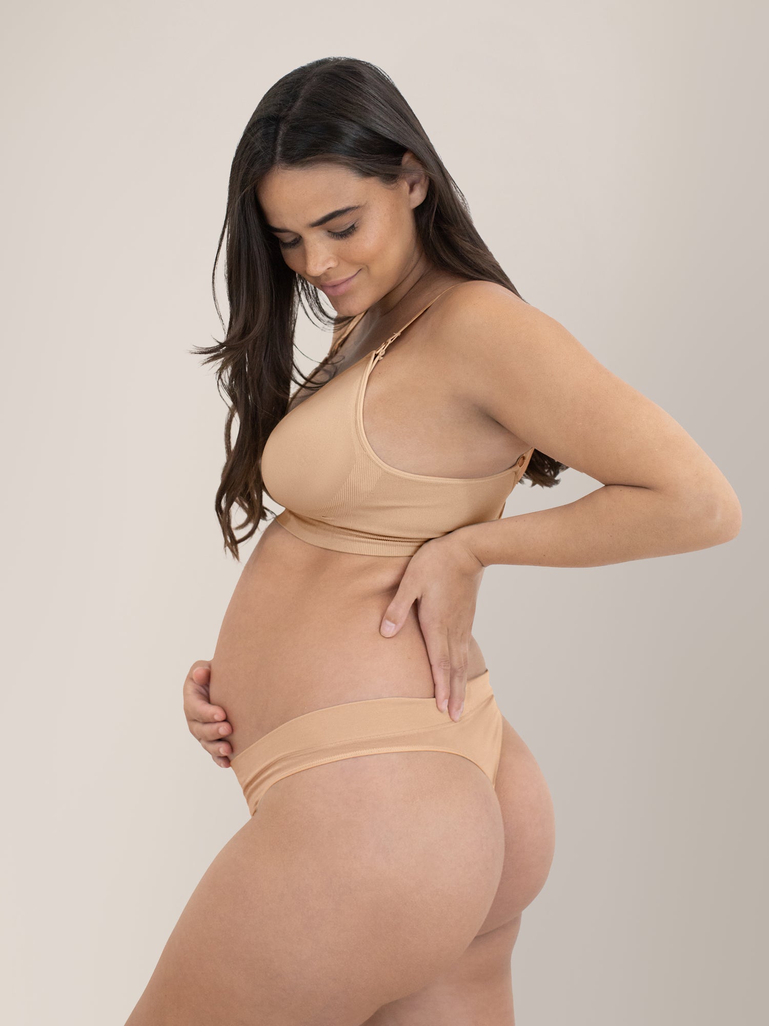 Pregnant model wearing the Bamboo Maternity & Postpartum Thong in Beige with her hand on her stomach and the middle of her back.