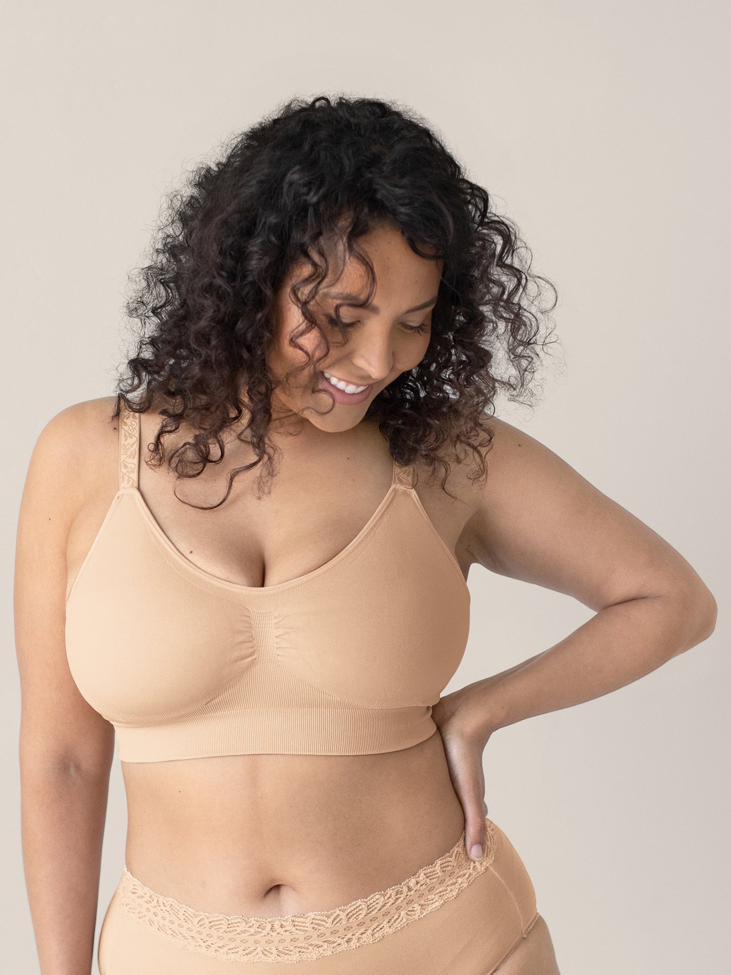 Model wearing the Nellie Sublime® Wireless Bra in Beige with her hand on her hip looking down at the bottom of the picture. @model_info:Sierra is wearing a Medium Busty.