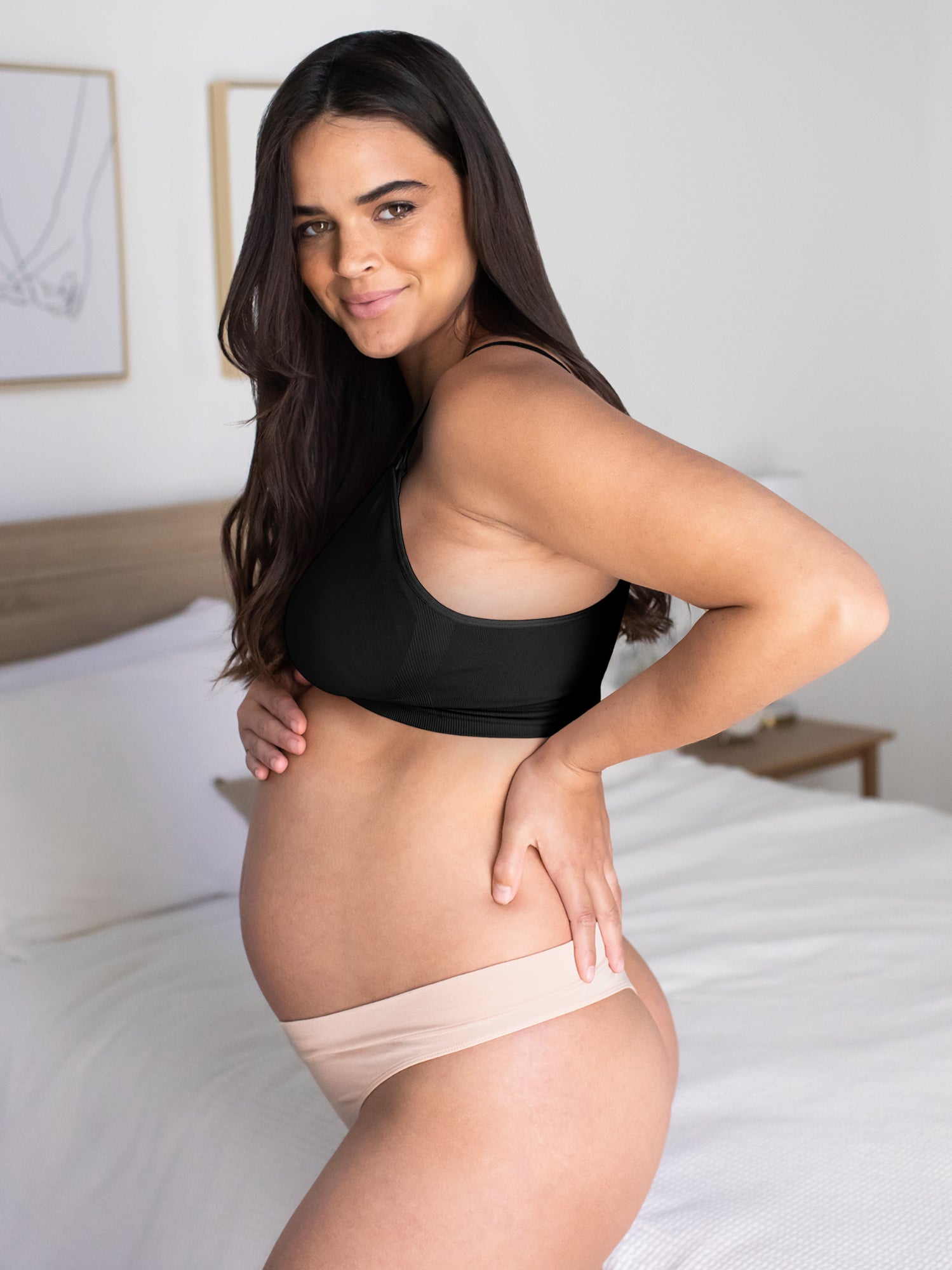 Side view of a model wearing the Bamboo Maternity & Postpartum Thong in Soft Pink  standing in front of the bed.