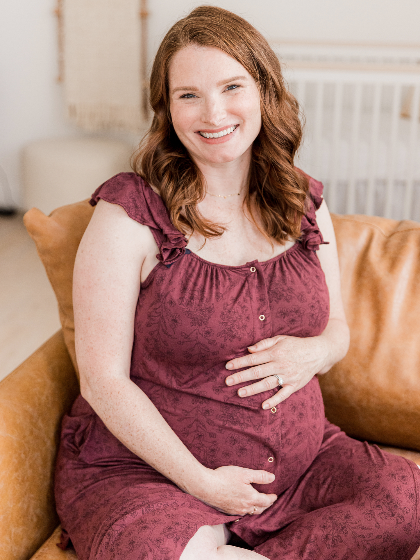 Pregnant model sitting on a couch while holding her stomach and wearing the Ruffle Strap Labor & Delivery Gown in Spice Floral