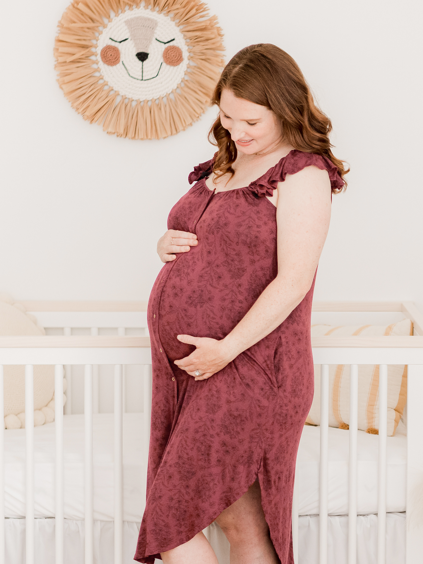 Pregnant model wearing the Ruffle Strap Labor & Delivery Gown in Spice Floral standing in front of a crib.