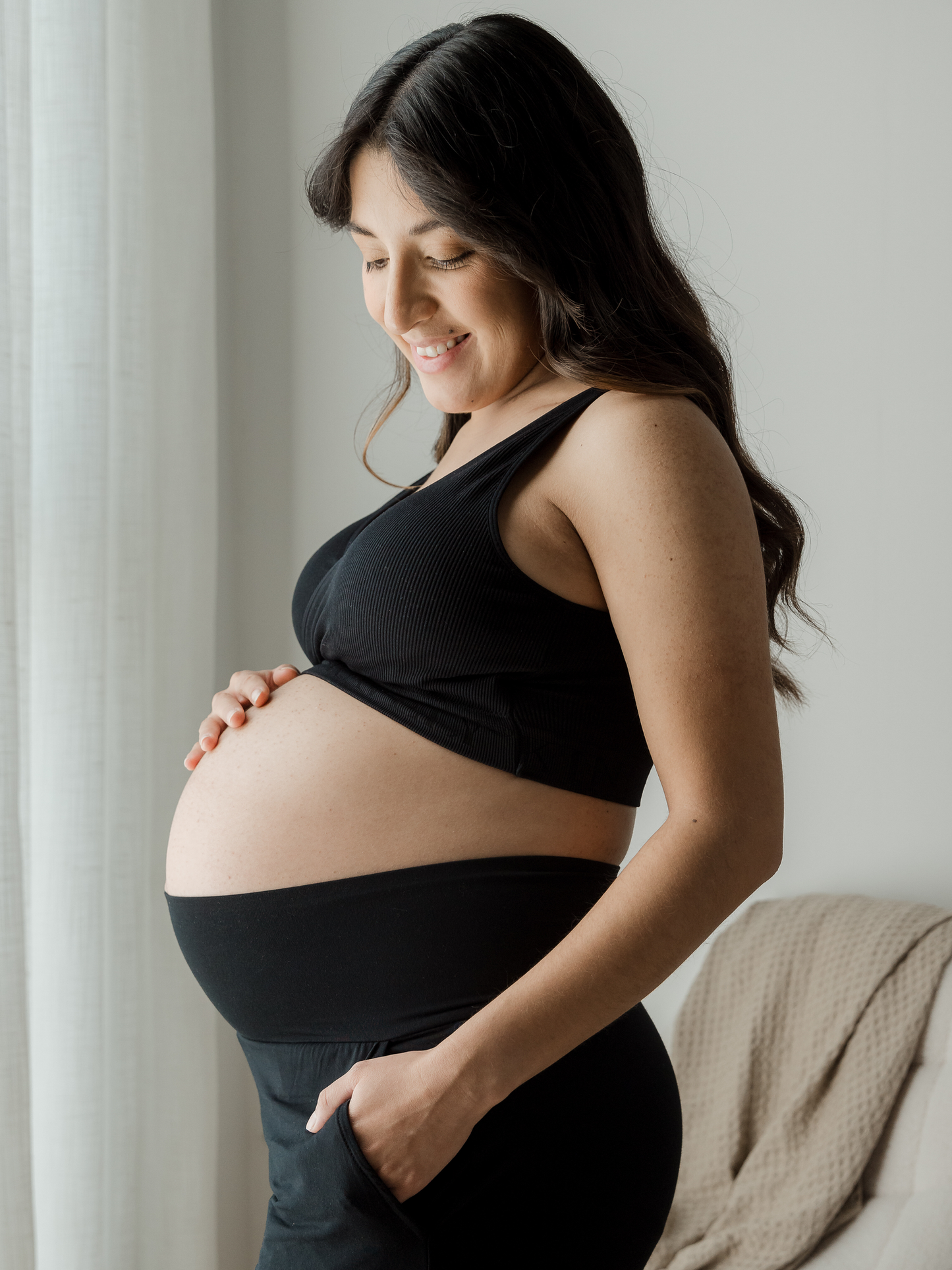 Pregnant model wearing the Sublime® Adjustable Crossover Nursing & Lounge Bra in Black holding her belly with her hand in her pocket. @model_info:Patty is wearing a Medium Busty.