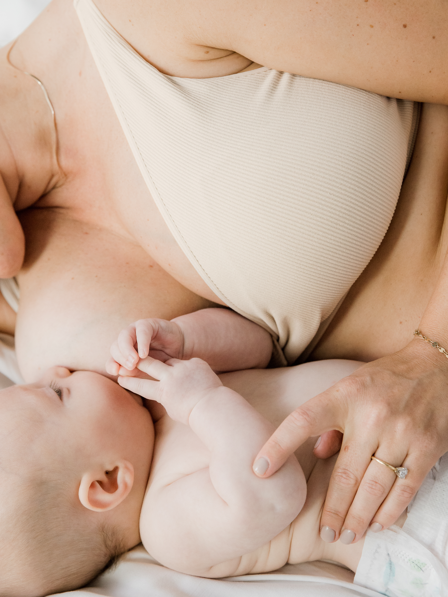 Model laying on a bed with her breastfeeding baby wearing the Sublime® Adjustable Crossover Nursing & Lounge Bra in Stone.