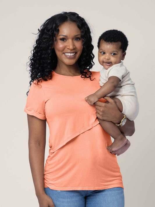 Model wearing the Everyday Asymmetrical Nursing T-shirt in Vintage Coral, holding her four month old baby @model_info:Rashé is wearing a Small.