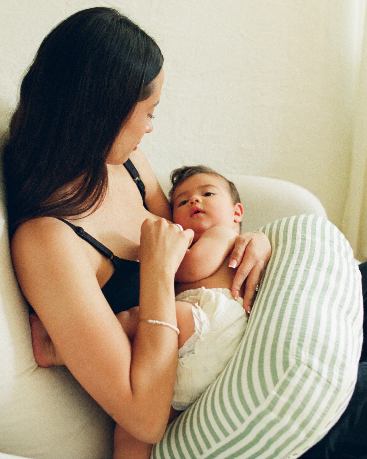 A mother breastfeeding baby on an Organic Muslin Nursing Pillow Cover in Sage Stripe in a cozy chair in a sun-filled room.