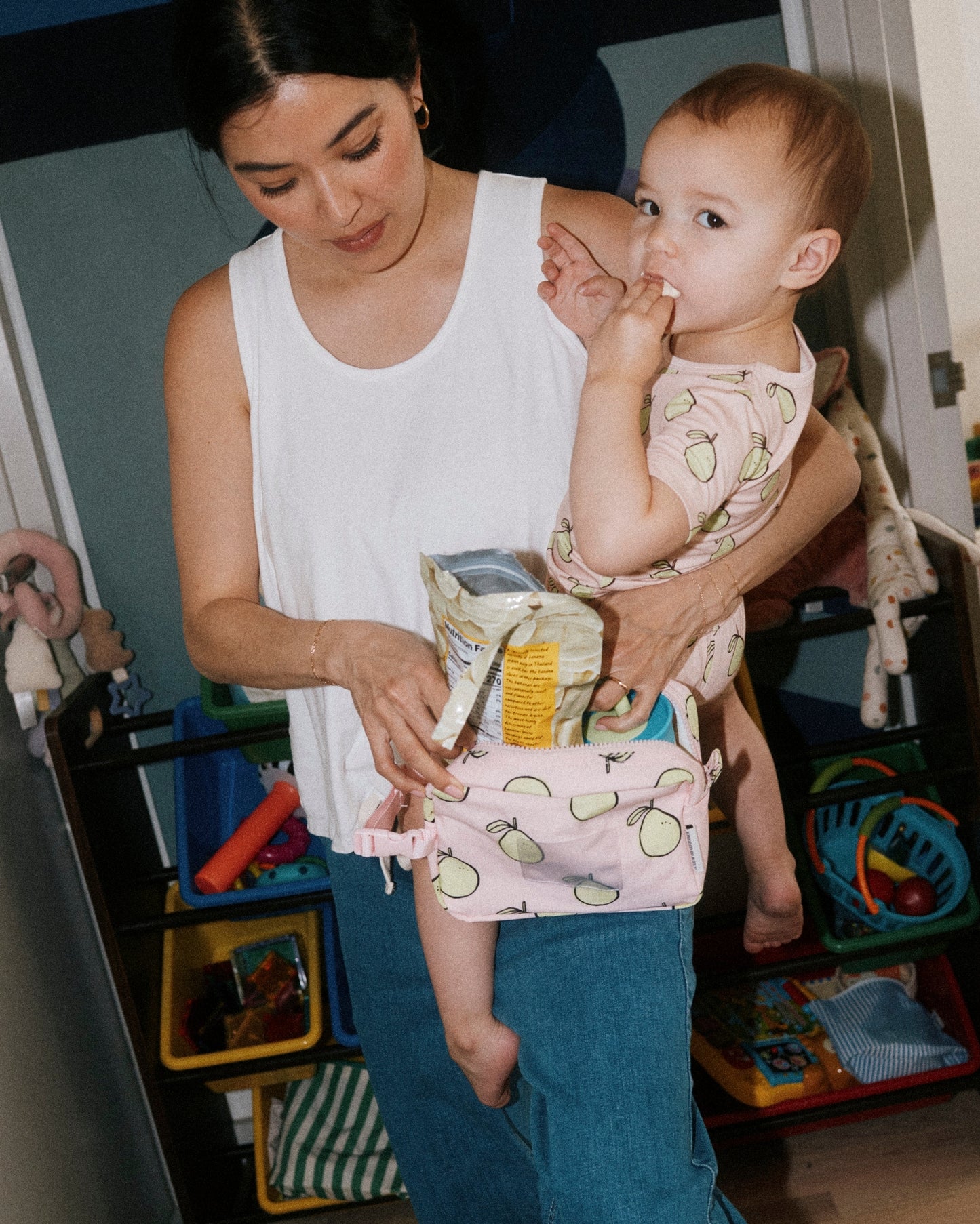 Woman holding toddler wearing Bamboo Short Sleeve & Shorts Set in Pink Pomelo while eating snacks from Mini Cooler Bag in Pink Pomelo. Woman is wearing Modal Signature Nursing Tank in White and Wide Leg Maternity & Postpartum Jean in Medium Wash, against a background of playroom full of toys and toy organizers.