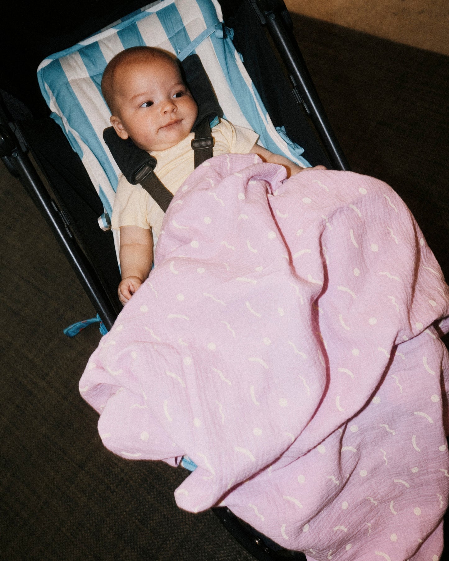 Baby sitting in stroller, on the Stroller Party Seat Liner in Blue Wide Stripe, covered by Organic Muslin Swaddle by Lilac Squiggles and wearing Honey Onesie from the Baby Onesie Two-Pack in Honey Dandelion, on a hallway carpet background.
