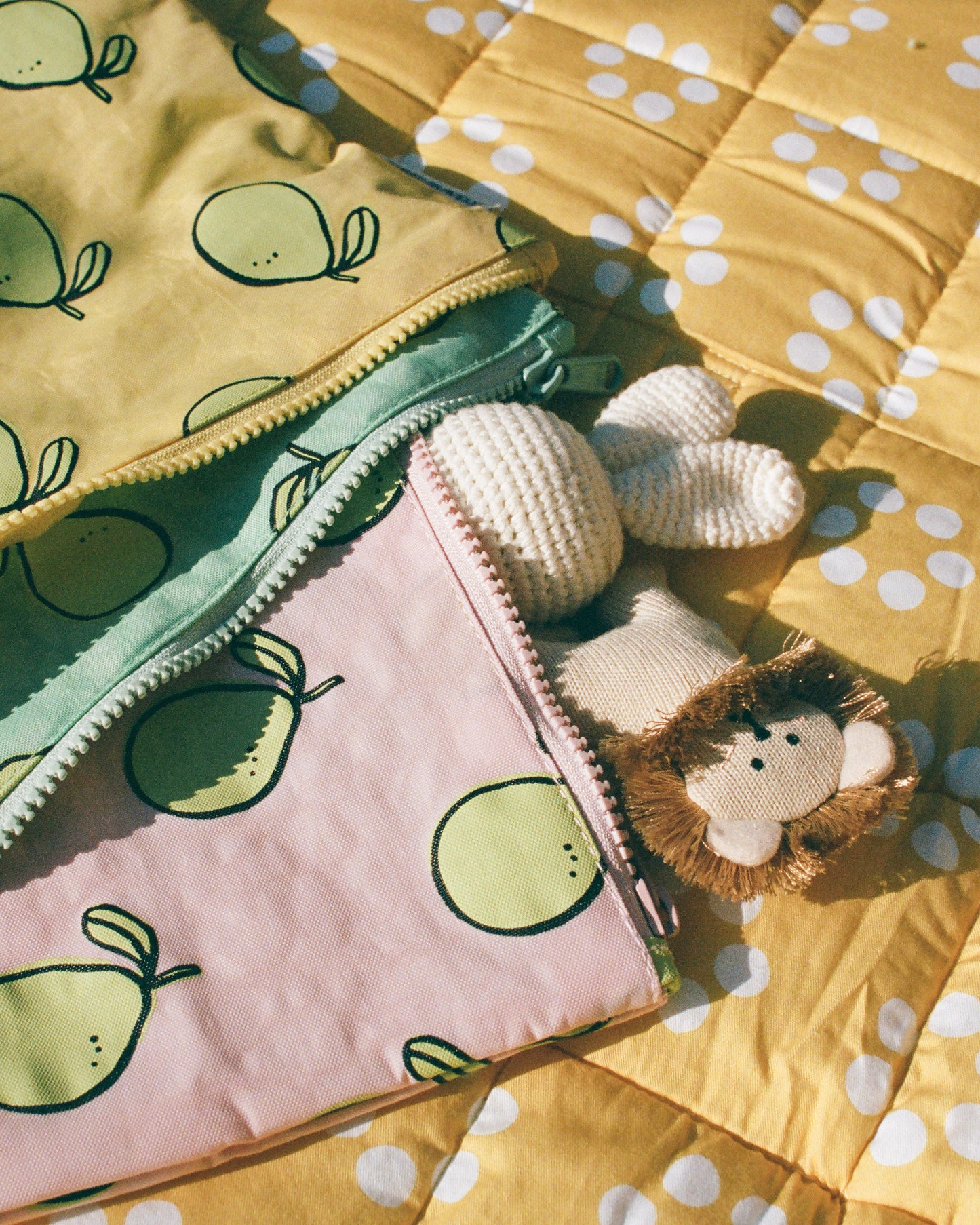 Zipper Pouch Three-Piece Set in Pomelo holding a bunny lovey and lion chewy toy, laying on the Inside/Outside Play Mat in Sunshine Dots in late afternoon sun.