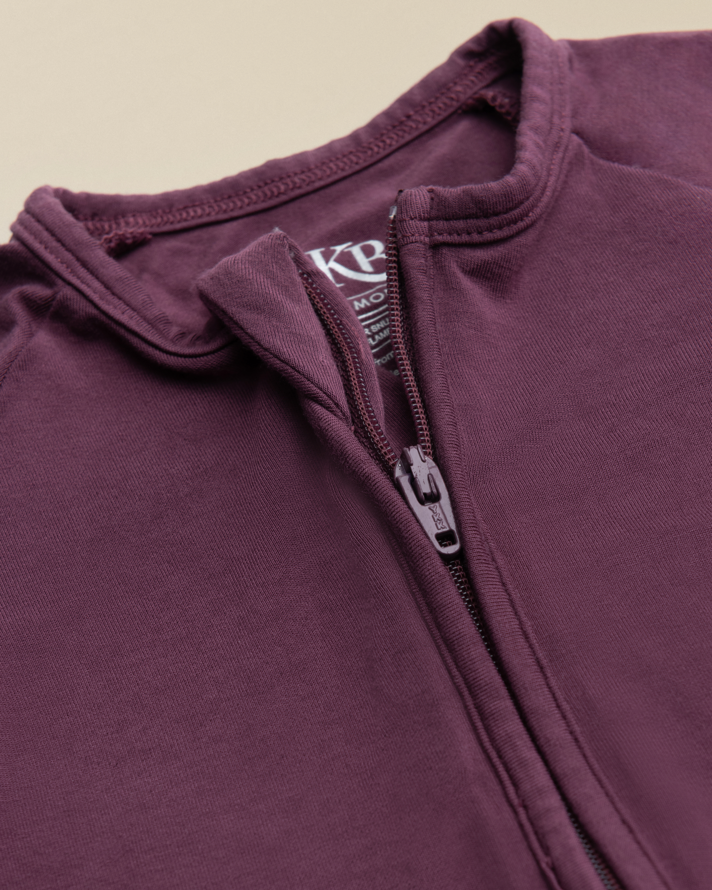 Close-up of Bamboo Baby Pajamas in Burgundy Plum with zipper and brand logo on a beige background