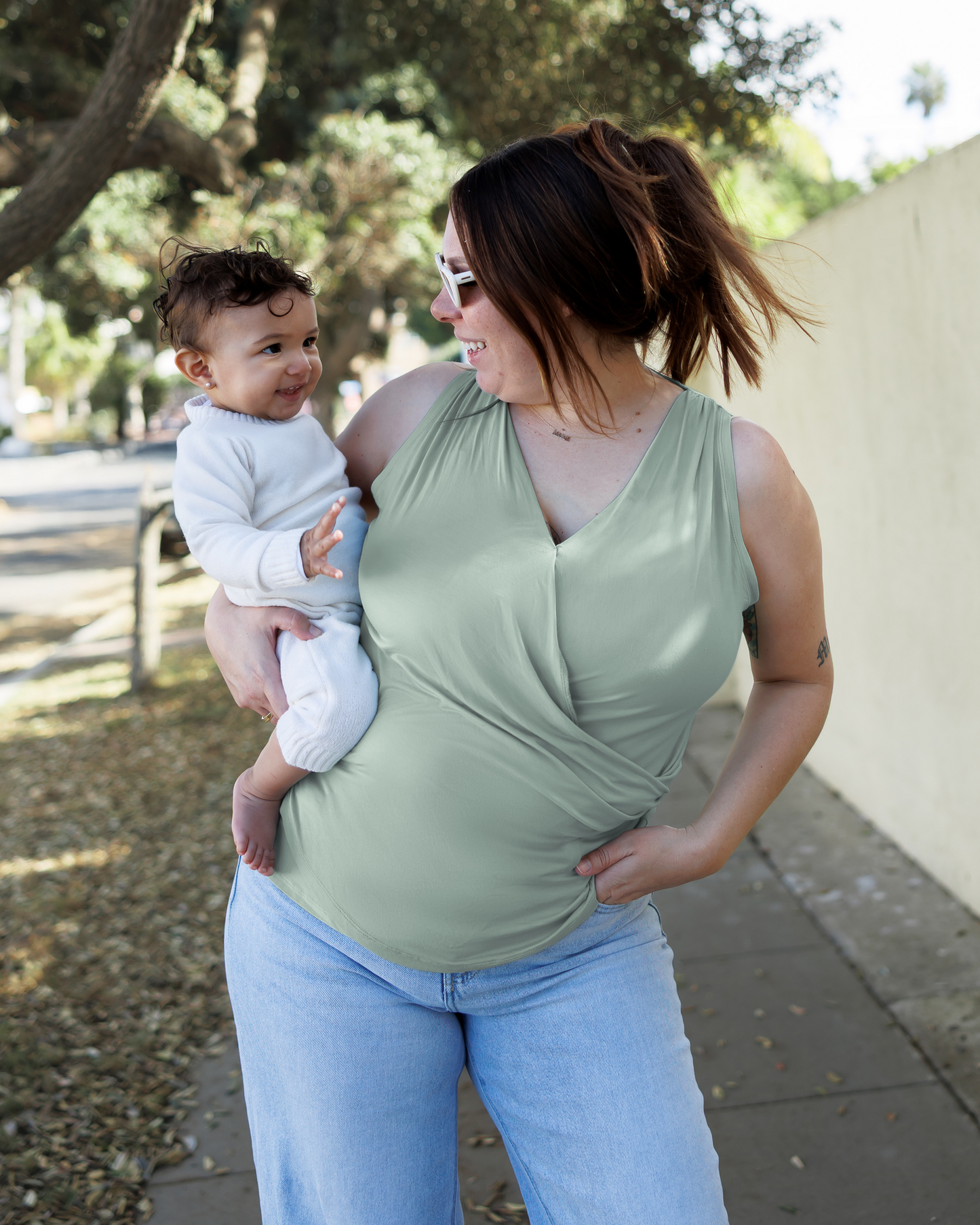 Front view of model holding baby and wearing the Bamboo Draped Nursing Tank in Aqua Mist, paired with jeans @model_info:Theresa is wearing a Large.