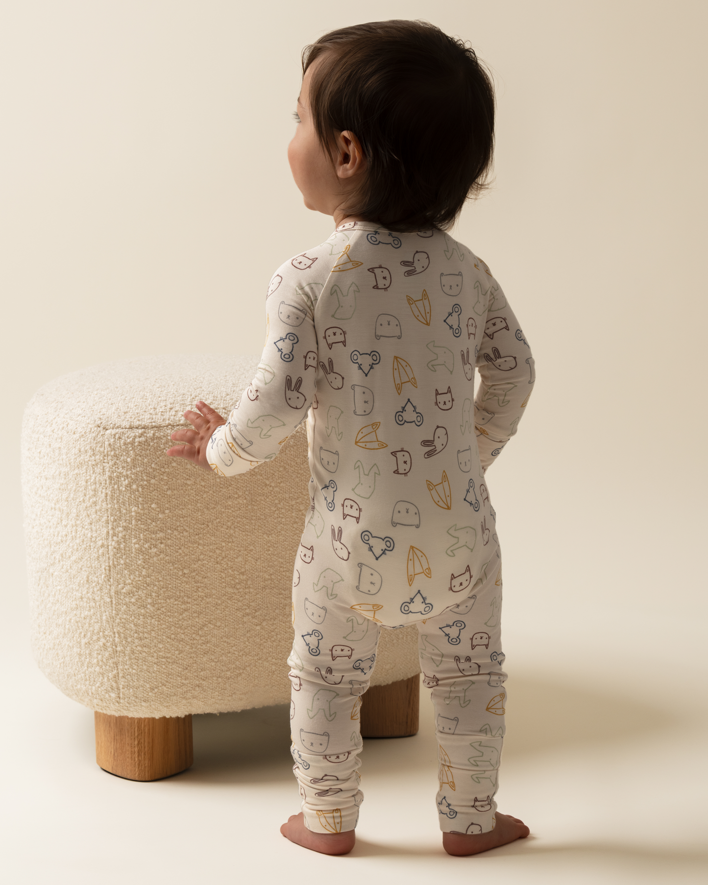 Child wearing Bamboo Baby Pajamas in Critter Crew standing next to a textured ottoman.
