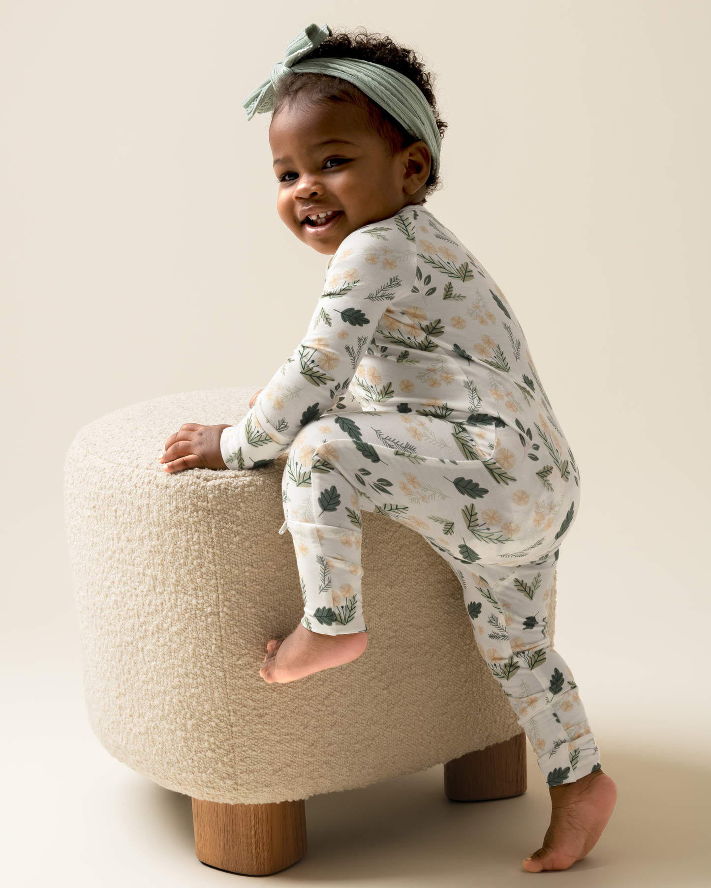 Child wearing Bamboo Baby Pajamas in Honey Dandelion sitting on a small ottoman against a plain background