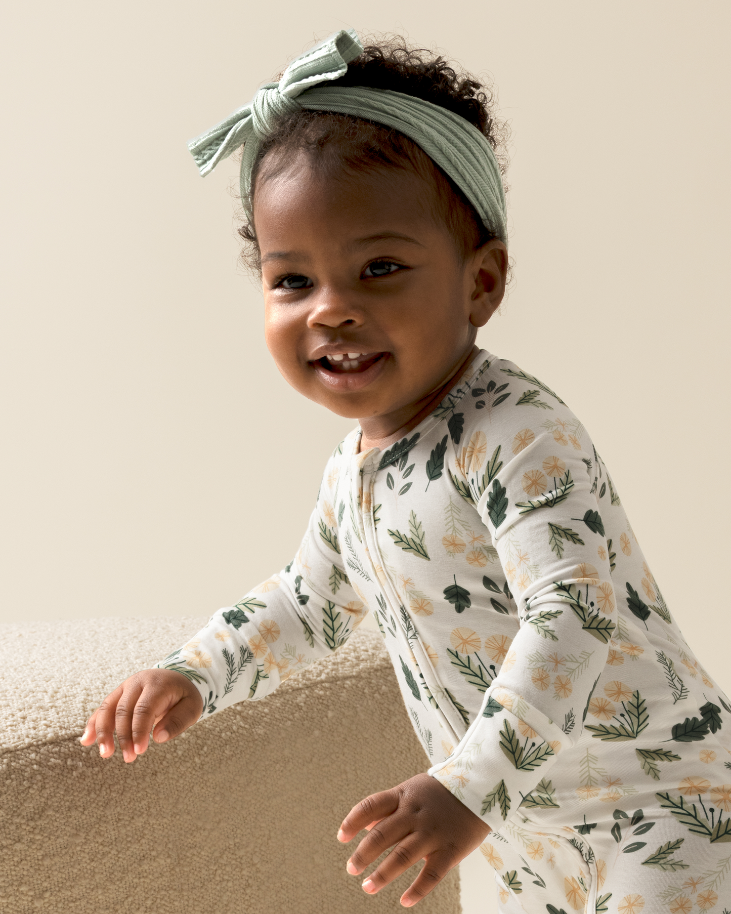 Baby wearing Bamboo Baby Pajamas in Honey Dandelion with a green headband against a beige wall