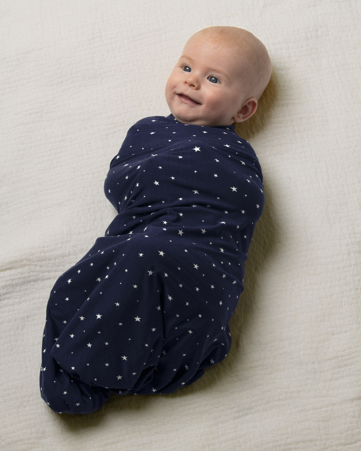 Baby wrapped in Bamboo Swaddle Blanket in Navy Stars on a beige surface@model_info:Danny is 3 months old.