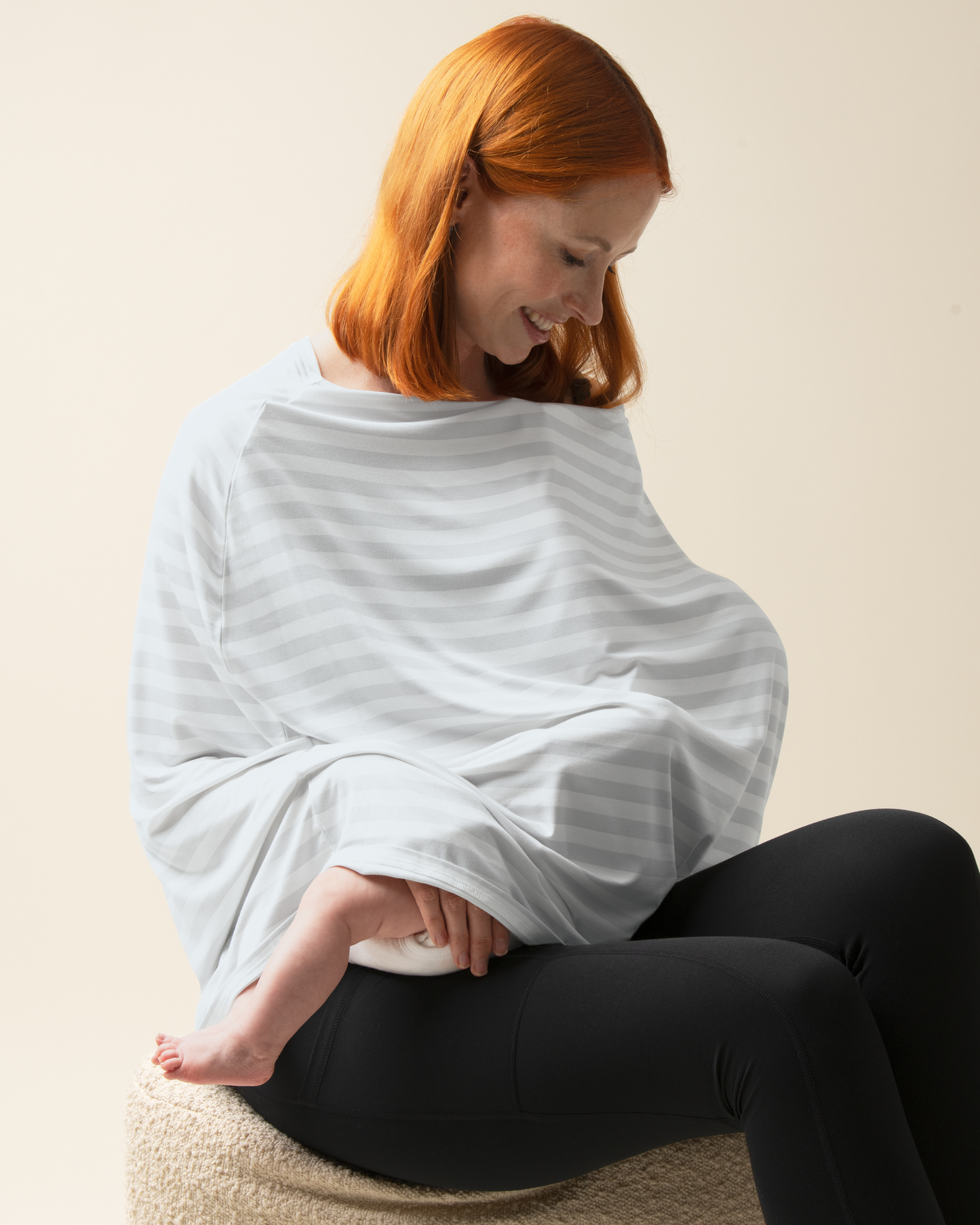 Woman holding a baby wrapped in a light blue blanket against a beige background wearing Bamboo Multi-Use Nursing Cover in Grey Stripe