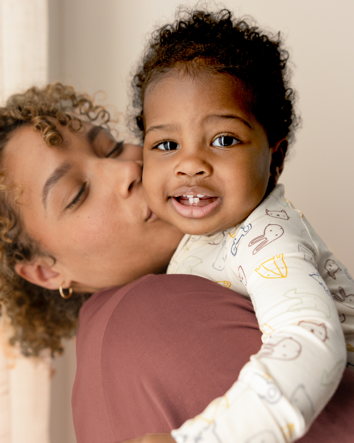 Woman kissing a baby on the cheek, with a neutral background. Baby wears Bamboo Baby Pajamas in Critter Crew@model_info:Harper is wearing 3-6 months.