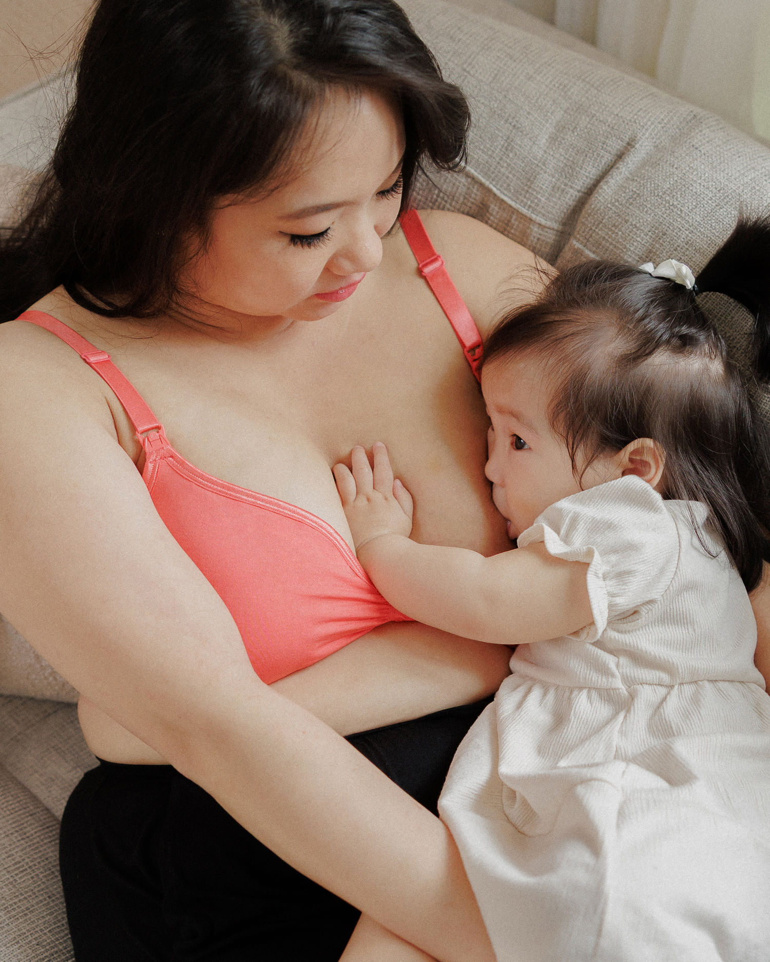 Model lounging on sofa cradling nursing daughter and wearing the Tatum Seamless Maternity & Nursing Bralette in Coral Sunset