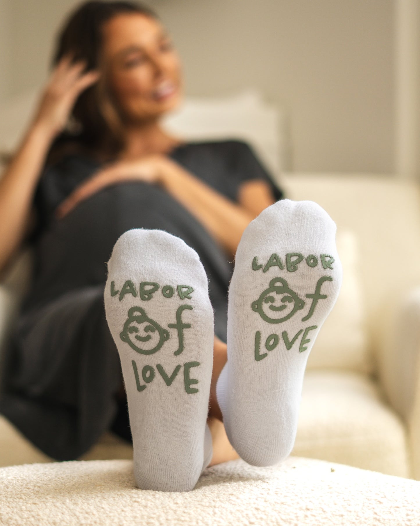 Model sitting on chair with feet on ottoman wearing Labor of Love Labor & Delivery Socks in sage