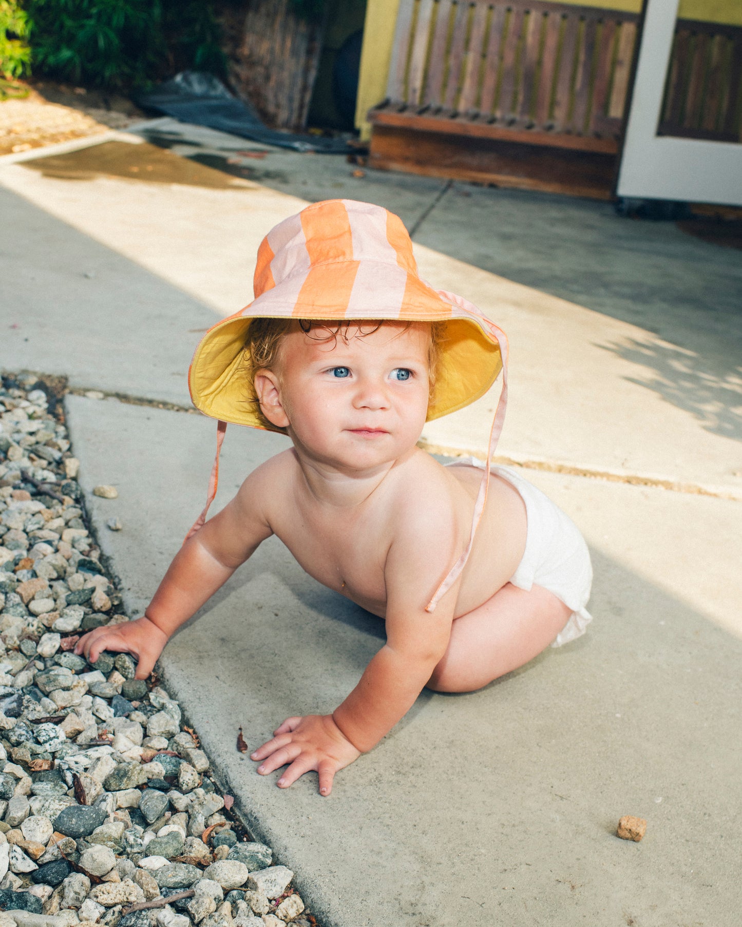 Baby is crawling on back patio and wearing Big Reversible Sun Hat in Pink Wide Stripe.