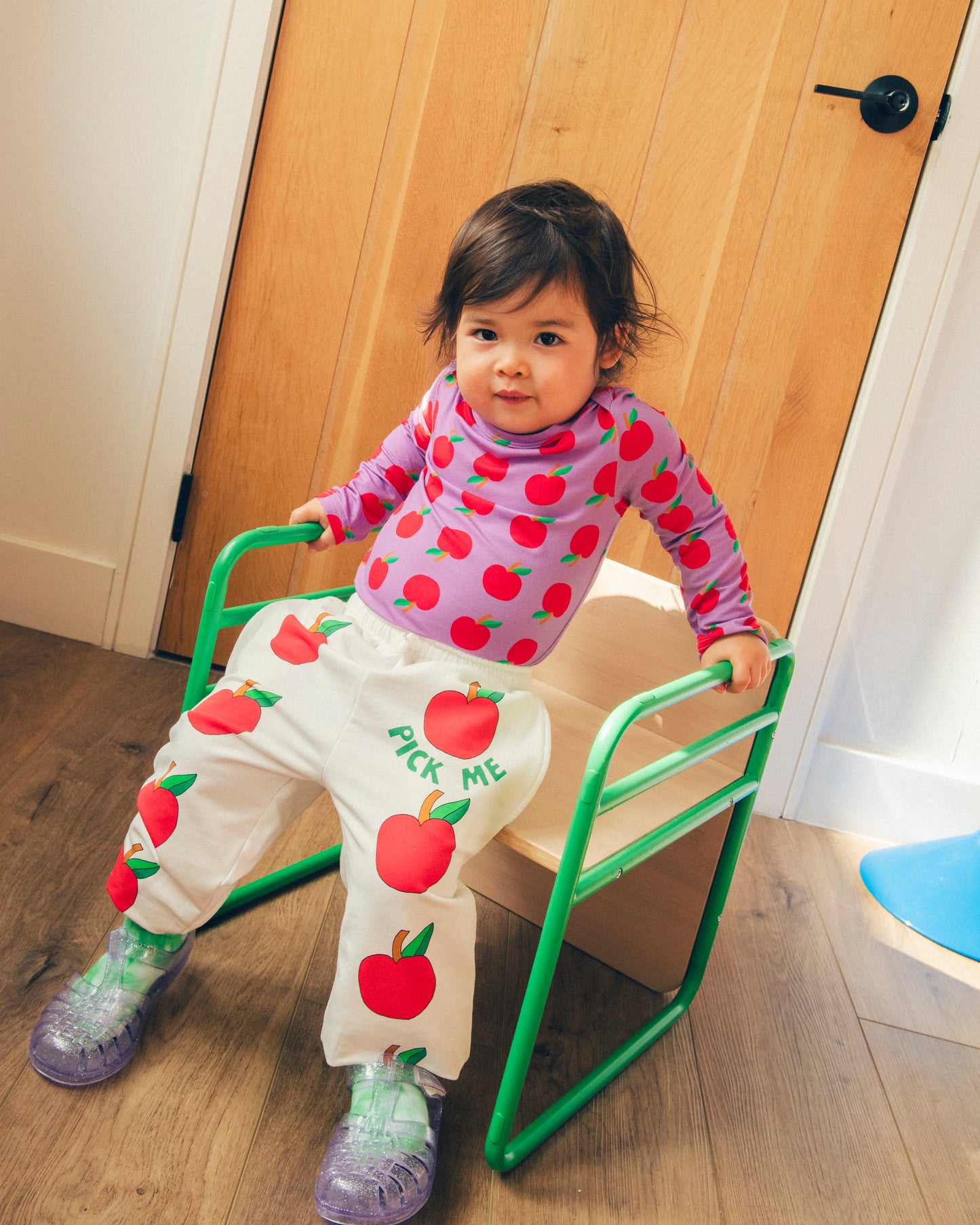 Model is wearing Purple Apple Onesie of the Long Sleeve Bamboo Onesie Two-Pack in Apple pattern and French Terry Jogger in Ivory Apple while sitting down in a chair indoors.