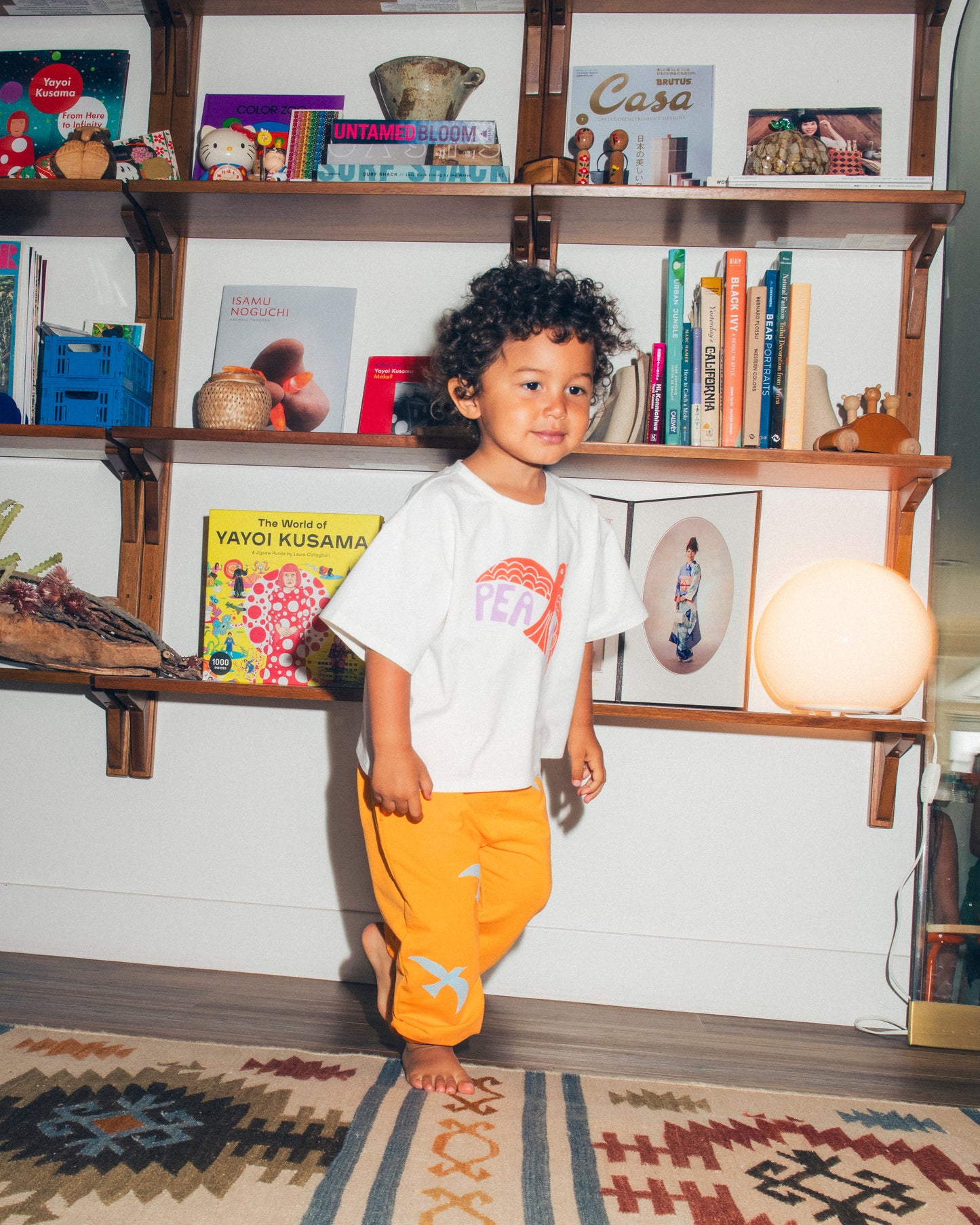 Model is wearing Graphic Tee in White Bird and French Terry Jogger in Orange Bird on a bookshelf background.@model_info:Kai is 2 and a half years old and wearing 3T.