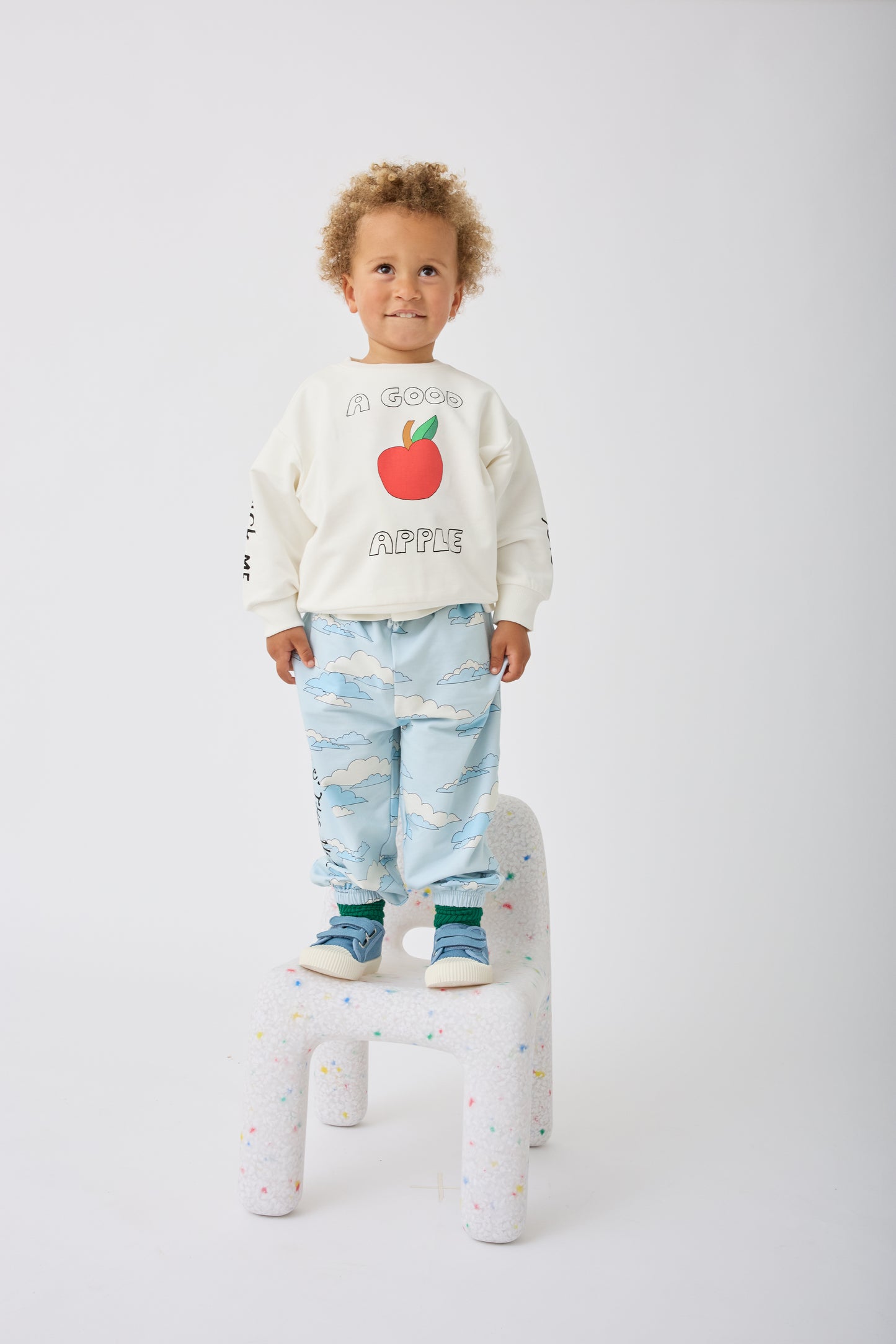 Model is wearing French Terry Jogger in Blue Clouds and Graphic Sweatshirt in Ivory Apple while standing on a chair on a gray background.