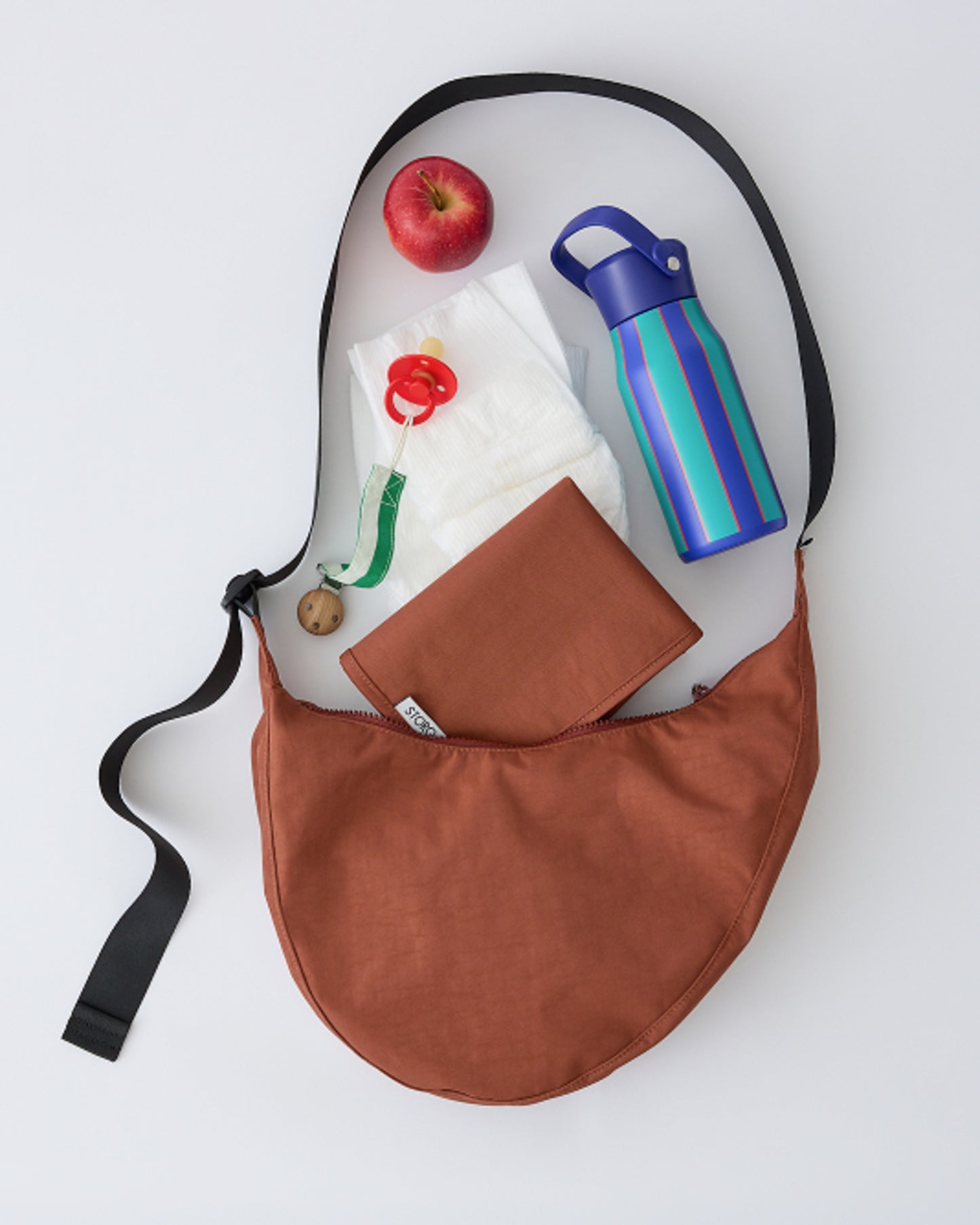 Flat lay of Half Moon Crossbody Bag in Cinnamon with snacks, water bottle, diaper, pacifier, and changing maton a beige background.