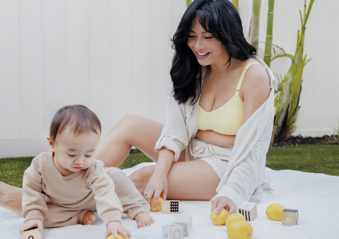 Model sitting on blanket on grass with baby wearing the Sublime® Hands-Free Pumping & Nursing Bra in Light Lemon, paired with the Reese Oversized Linen Button Down Shirt and the Reese Linen Short in Natural