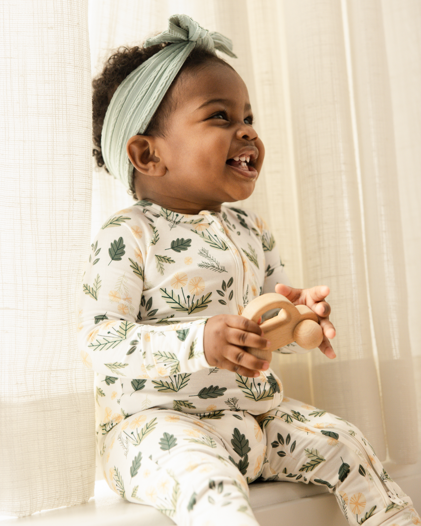 Child wearing Bamboo Baby Pajamas in Honey Dandelion holding a wooden toy, sitting against curtain @model_info:Harper is wearing 3-6 months.