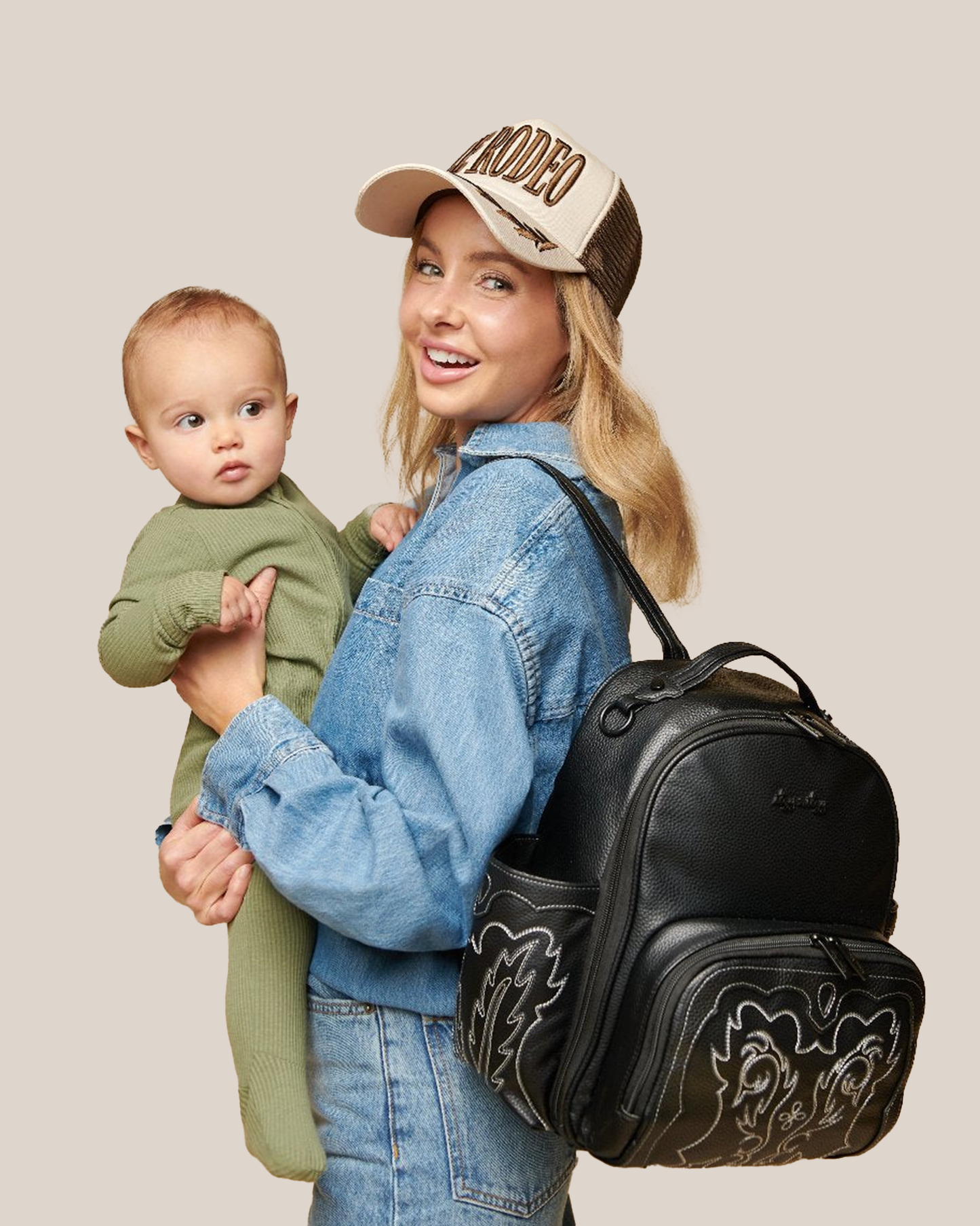 Model wearing the Itzy Ritzy - Nash Itzy Mini Plus™ Diaper Bag in Outlaw on one shoulder and carrying baby