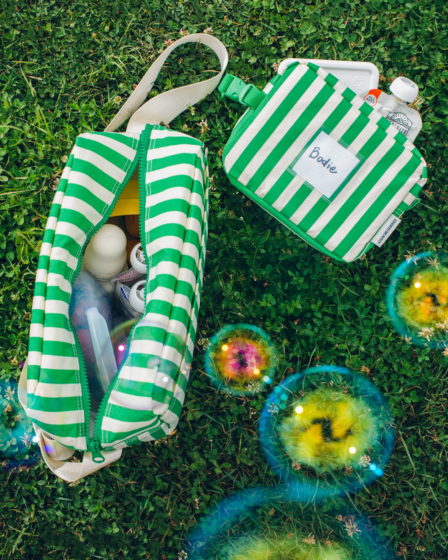 Jumbo Cooler Bag in Green Stripe and Mini Cooler Bag in Green Stripe sitting on grass, both filled with snacks and milk bottle, and bubbles floating above.