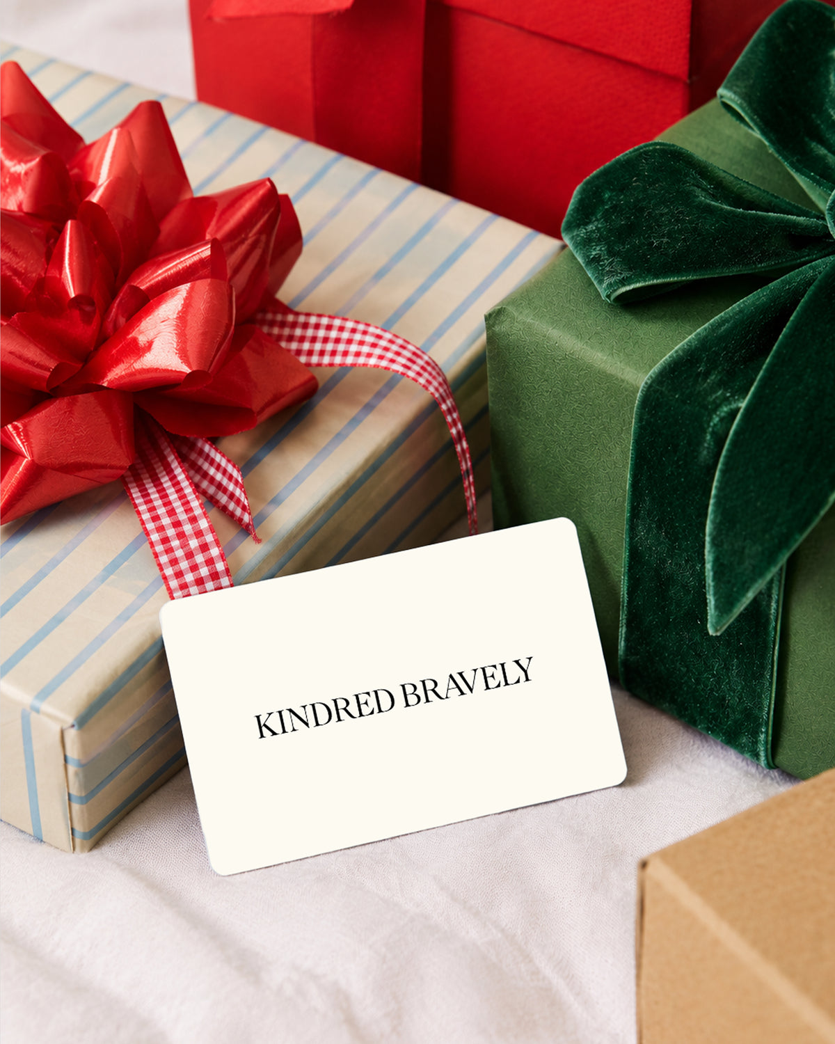 A white gift card with the text 'KINDRED BRAVELY' printed in black, styled on top of wrapped holiday gift boxes with red and green bows.