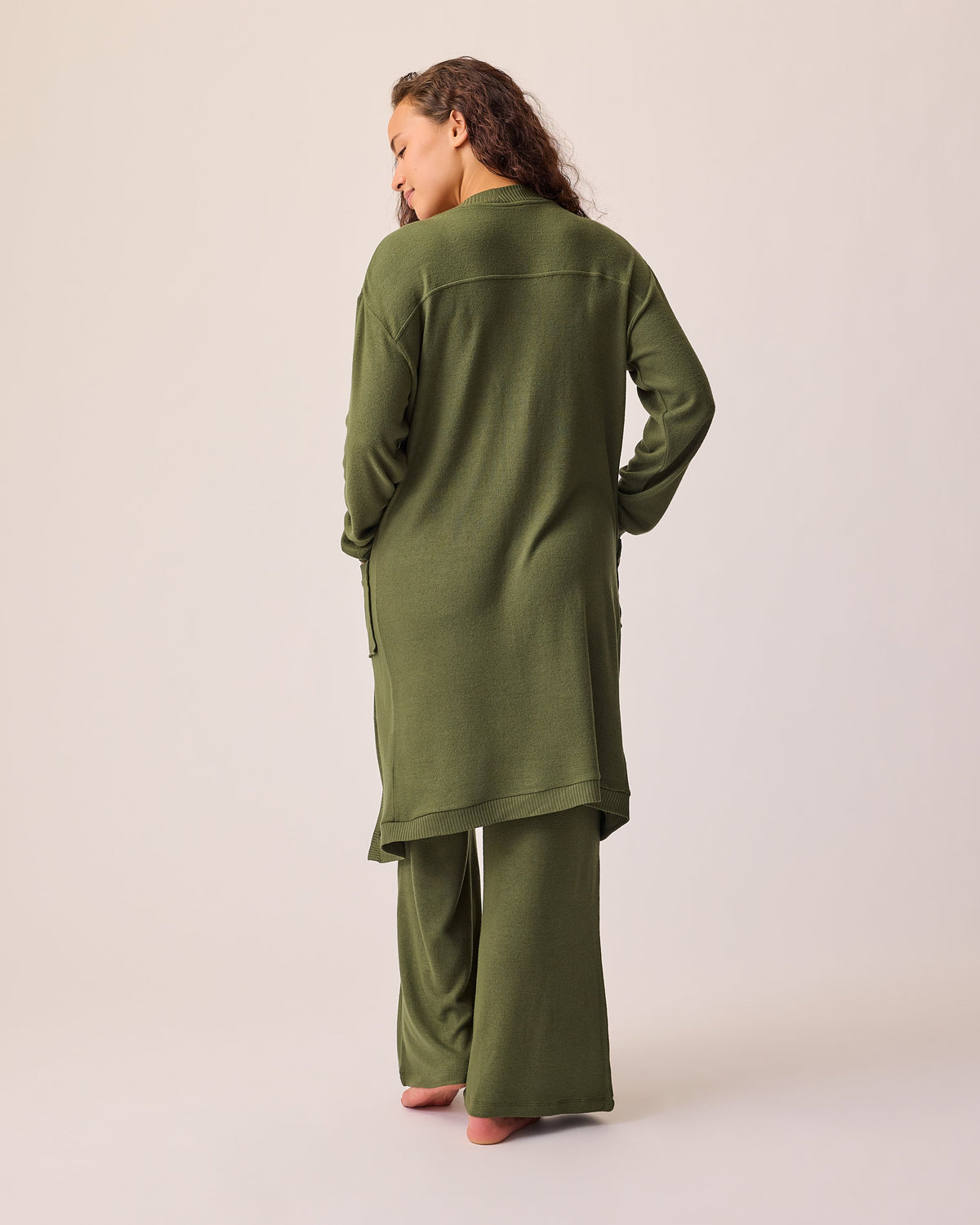 Back view of model wearing CozyKnit Long Cardigan in Deep Olive, with Simply Sublime® Nursing Bra in Deep Olive, and CozyKnit Wide Leg Lounge Pant in Deep Olive on a beige background.