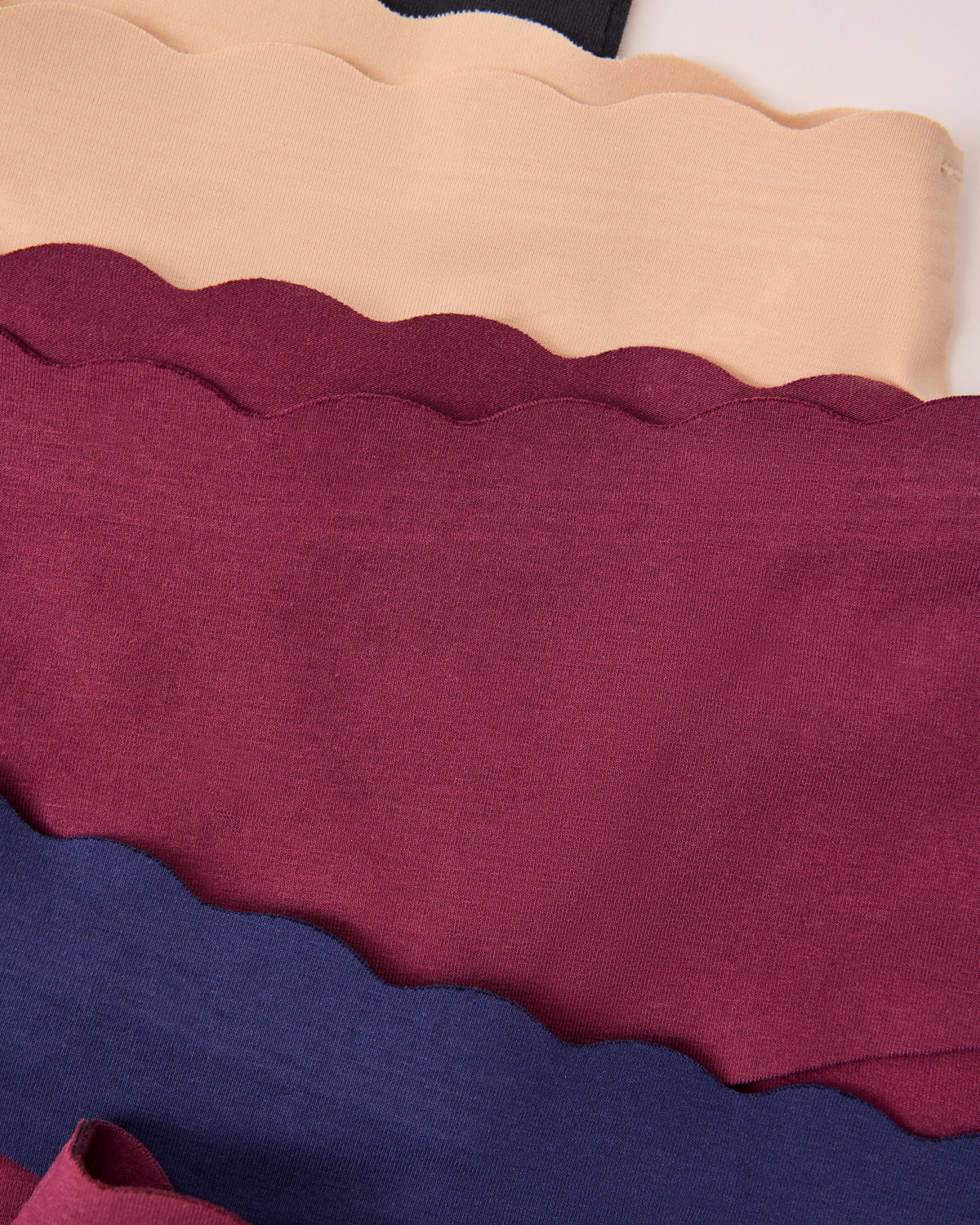 Four scalloped-edge fabric bands of the BareBond™ Cotton Maternity & Postpartum Underwear in Black, Sand, Garnet and Coastal Navy on a beige background.