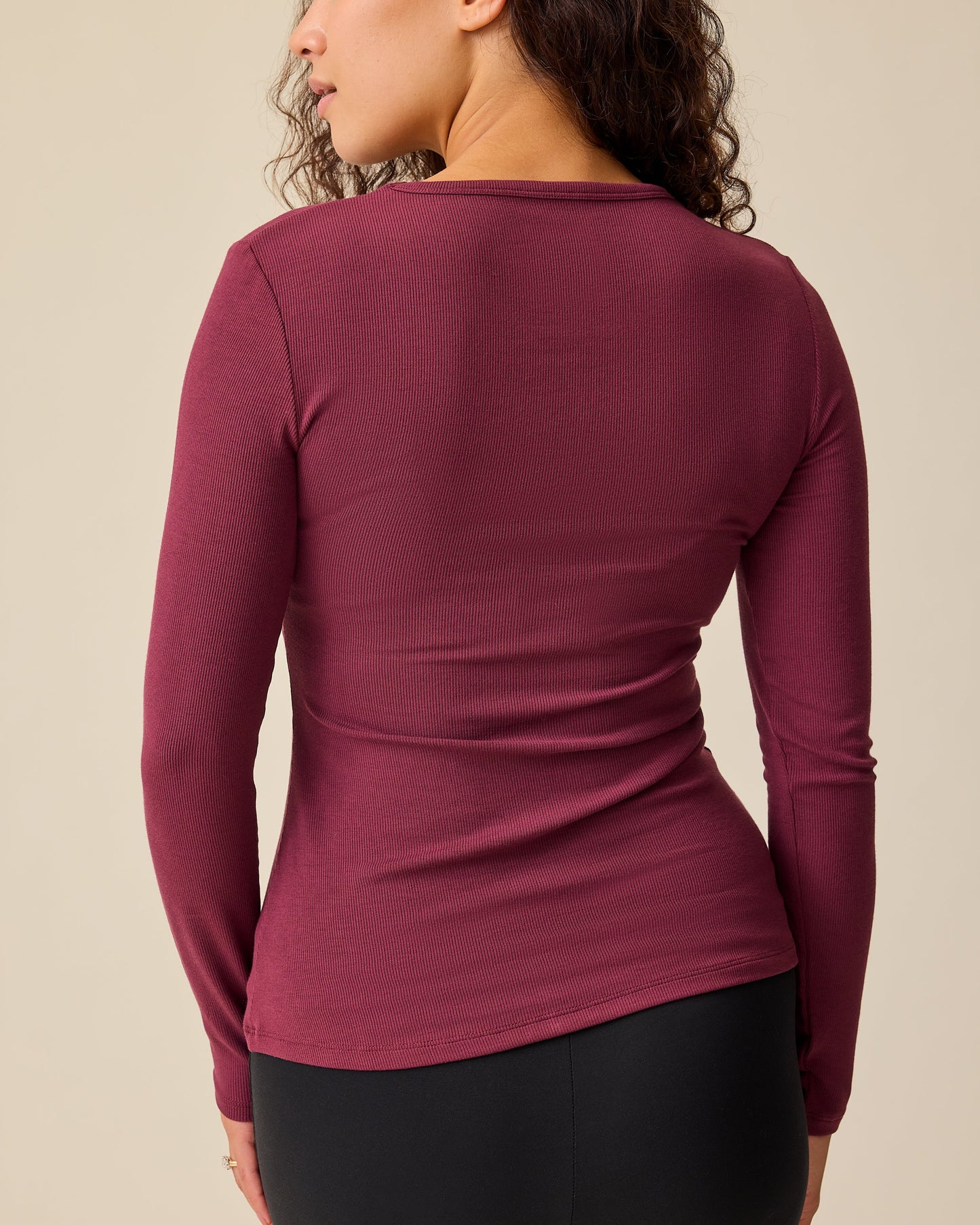 Back view of model wearing Ribbed Bamboo Long Sleeve Nursing T-shirt in Deep Burgundy with black pants, on a beige background.