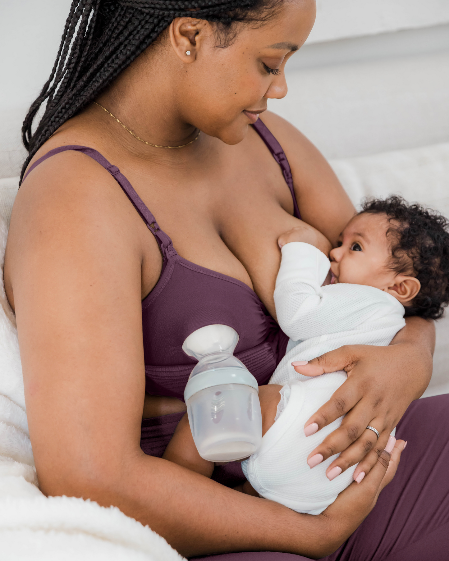 Model wearing the Sublime® Hands-Free Pumping & Nursing Bra in Burgundy Plum, with pump flange inserted on one side, and nursing baby on the other side