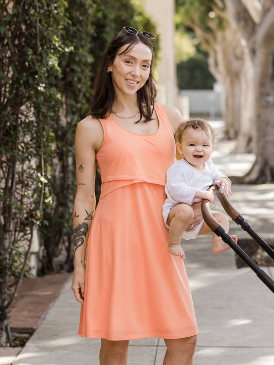 Model outside holding baby and wearing the Penelope Crossover Nursing Dress in vintage coral @model_info:Lyn is wearing a Small.