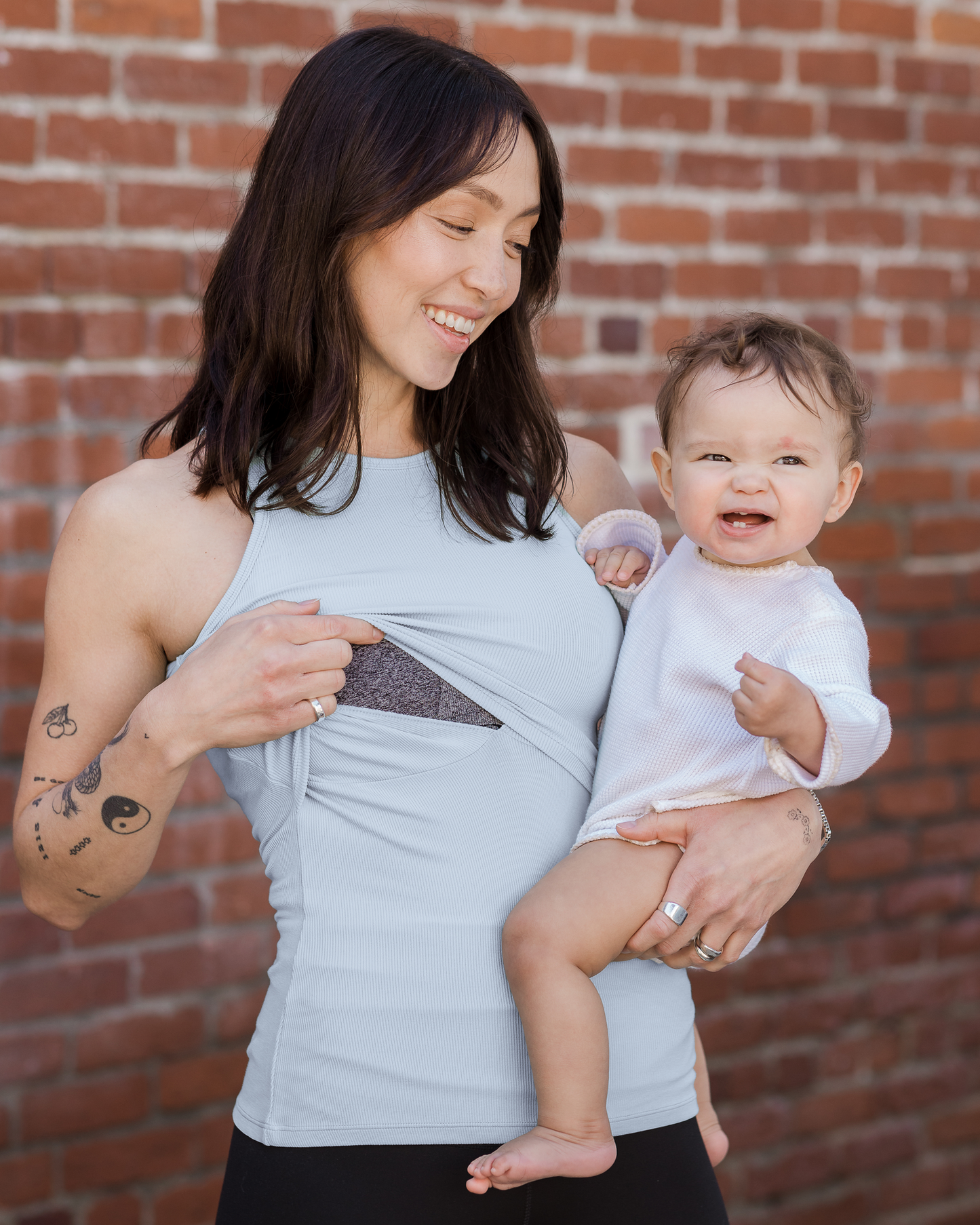 Model outside holding baby wearing the Ribbed Bamboo Racerback Nursing Tank in French Blue, lifting up top outer layer to show nursing access
