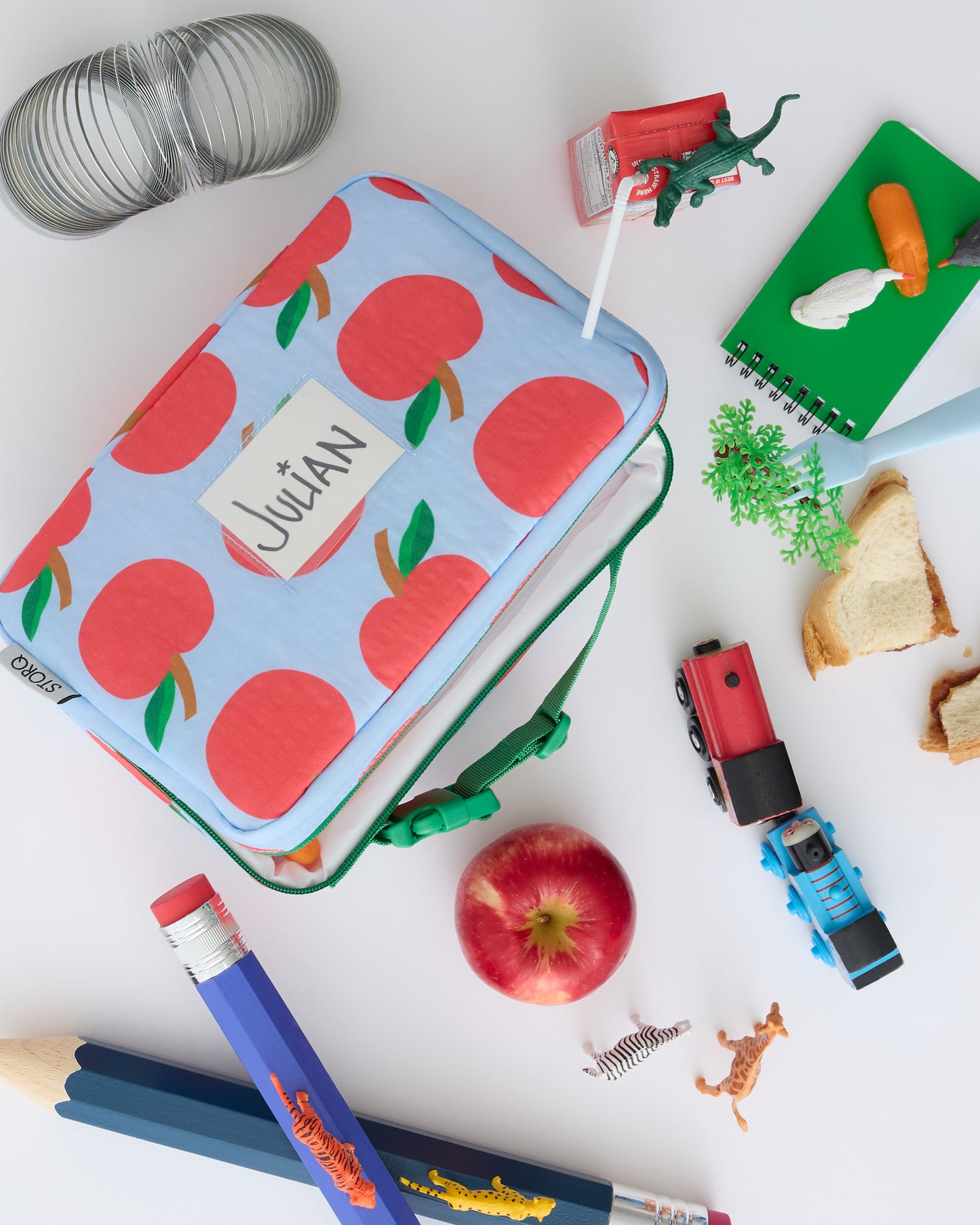 Flat lay of Cool Kid Lunch Box in Blue Apple with giant pencil, apple, notebooks, snacks, crayons, juice box, miniature animals and train toys on a beige background.