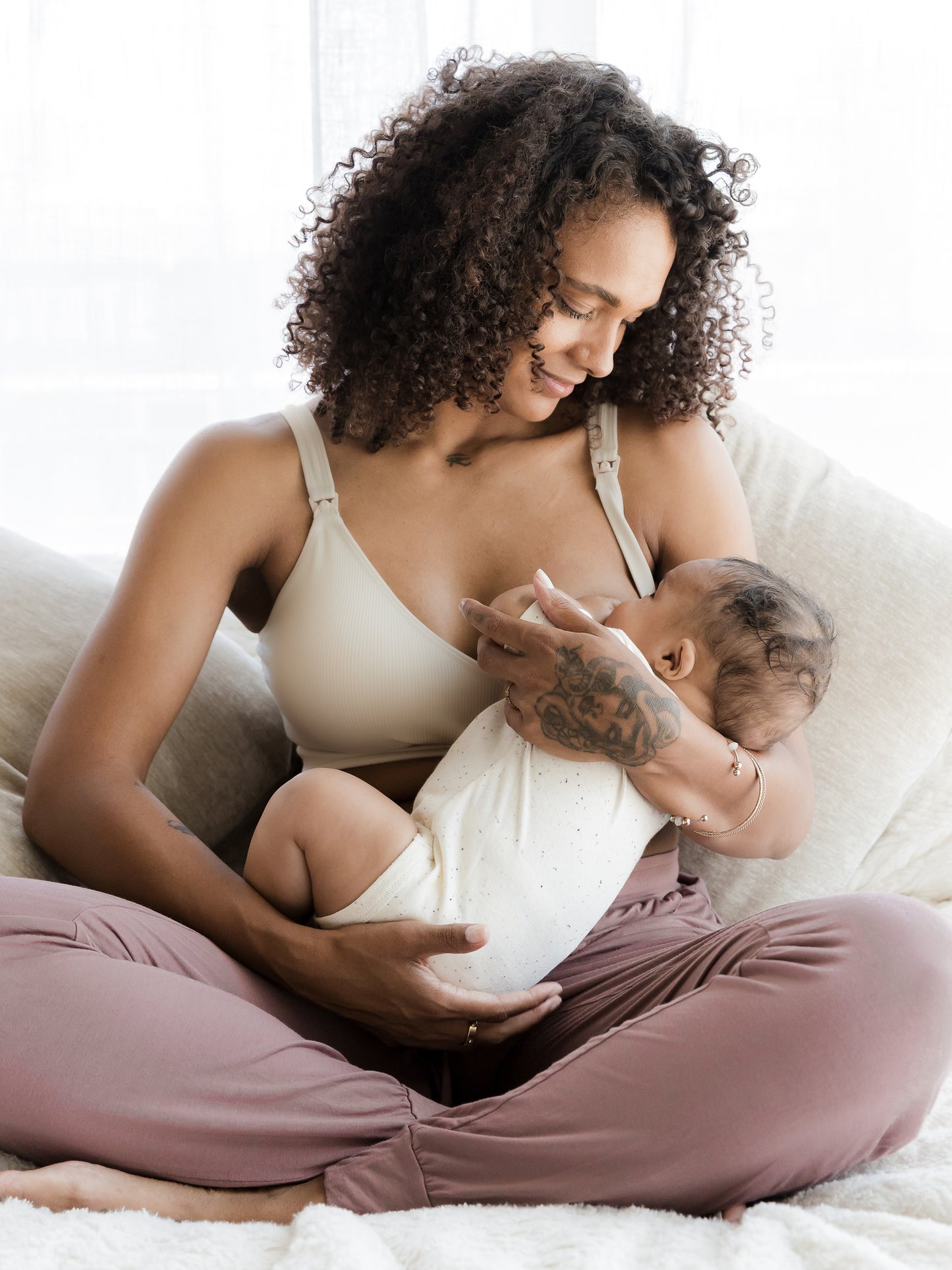 Model sitting on bed nursing baby and wearing the Mia Maternity & Nursing Lounge Bra in Stone. @model_info:Rudi is wearing a Small.