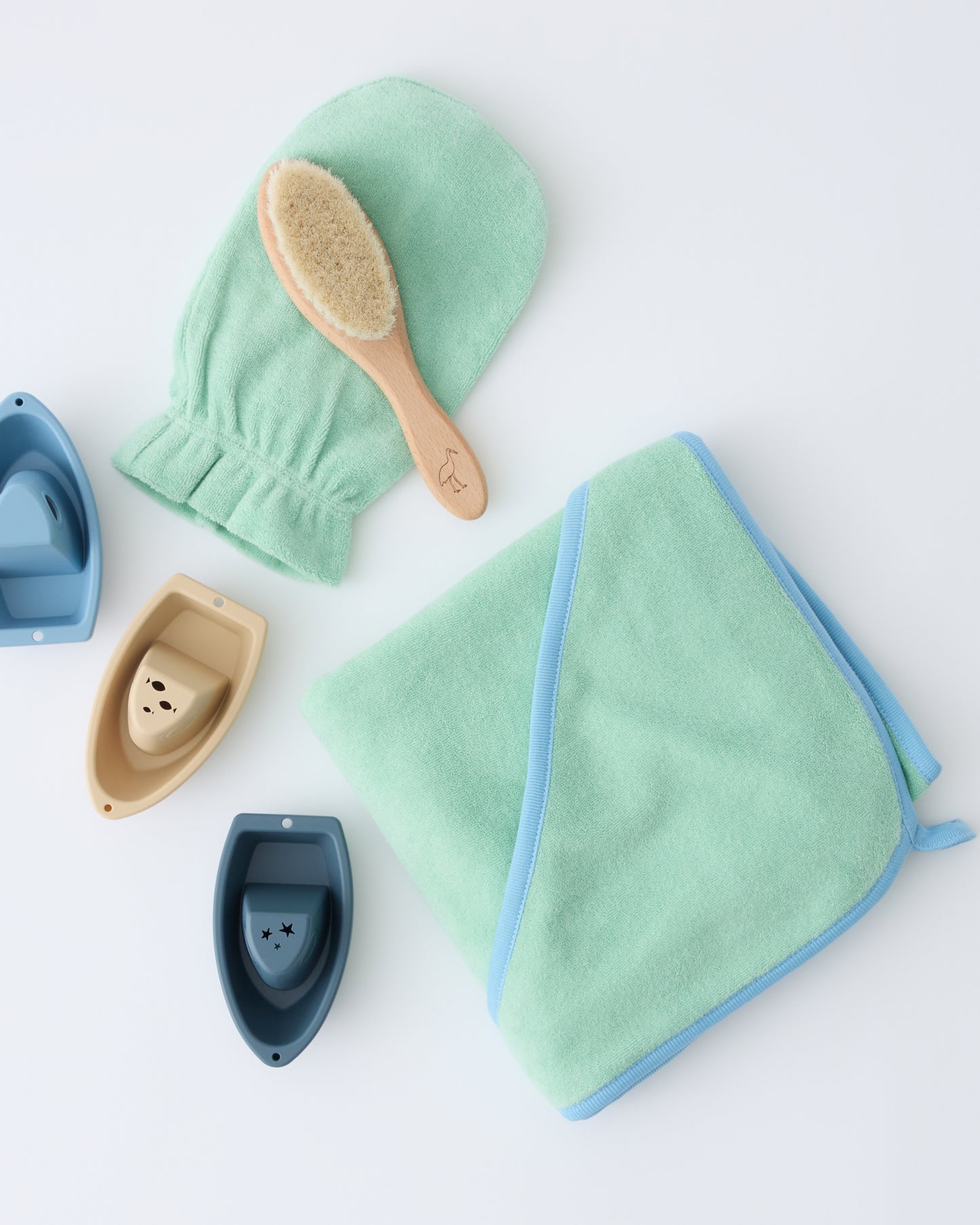 Newborn Bath Set in Mint, including hooded towel, hair brush and bath mitt, paired with three floating boat toys, on a beige background.