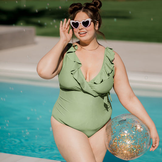 Model wearing the Ruffle Wrap Maternity & Nursing One Piece Swimsuit in Aloe standing by a pool with a beach ball on her hip. @model_info:Bailey is wearing a Large Busty.