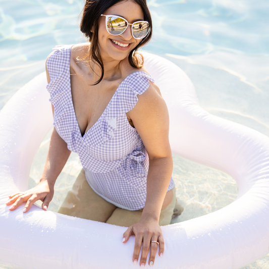 Model wearing the Ruffle Wrap Maternity & Nursing One Piece Swimsuit in Lavender Gingham with her hands on tan inner tube. @model_info:Nohely is wearing a Medium.